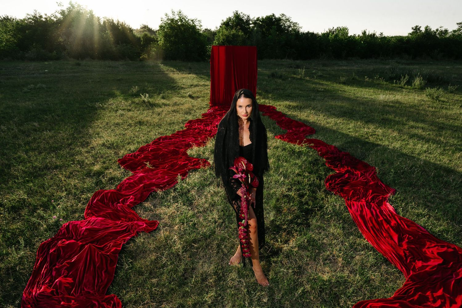 Woman in black outfit stands among flowing red fabric in a sunlit green field with a red door backdrop.