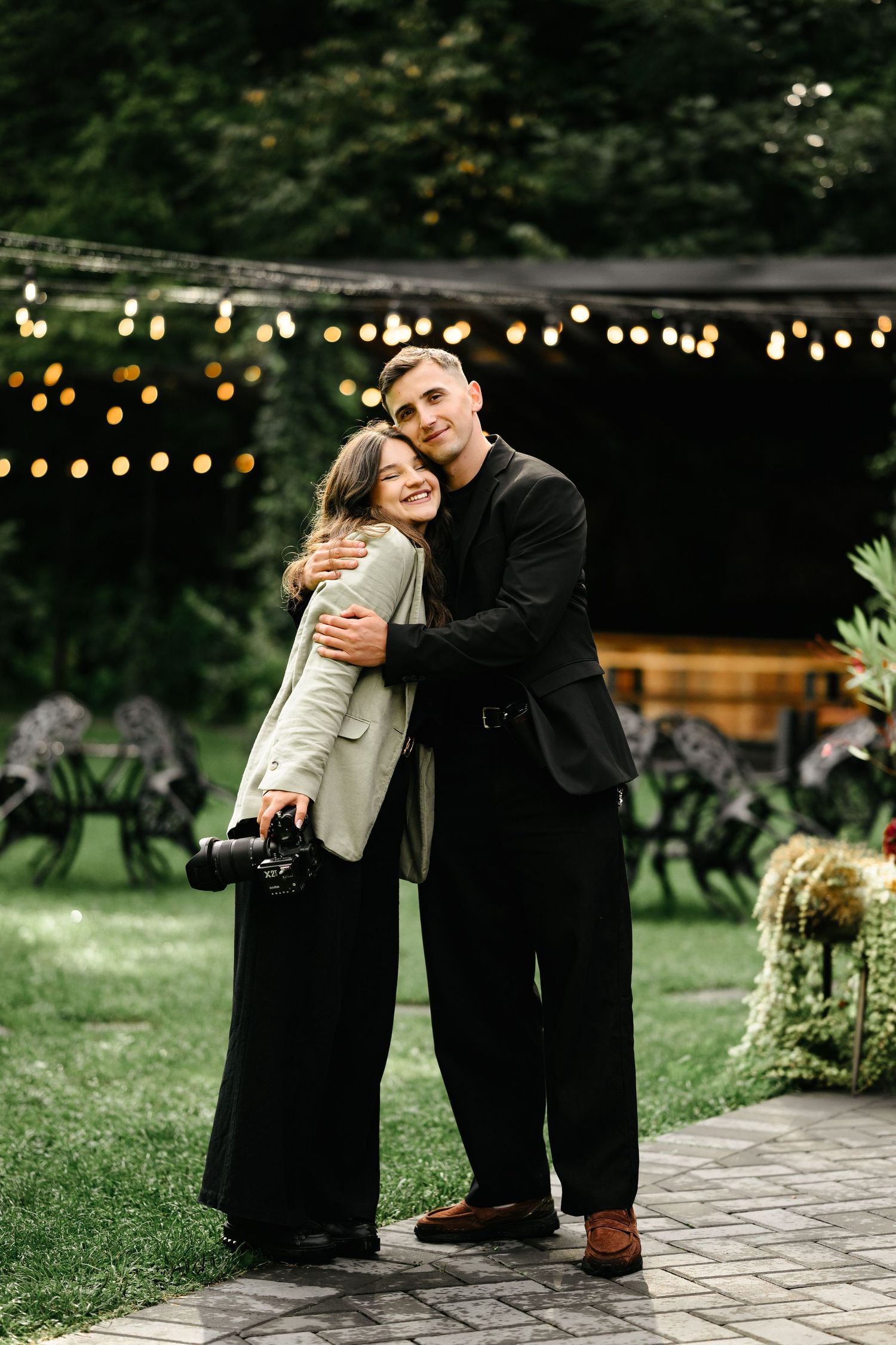 Couple embracing outdoors at evening garden event with string lights and floral decorations in background.