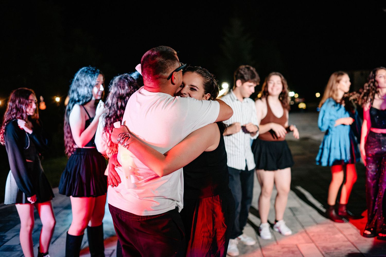 Couple sharing warm embrace at outdoor night celebration with group of people dancing in background.
