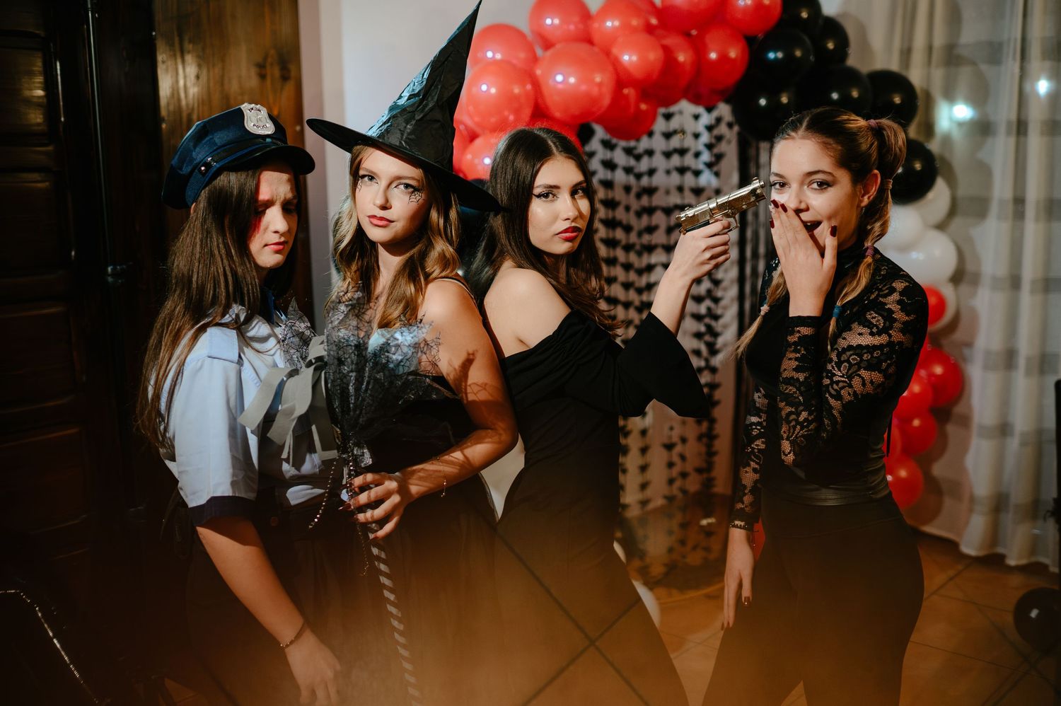 Four women in Halloween costumes including a witch and police officer pose by festive balloons.