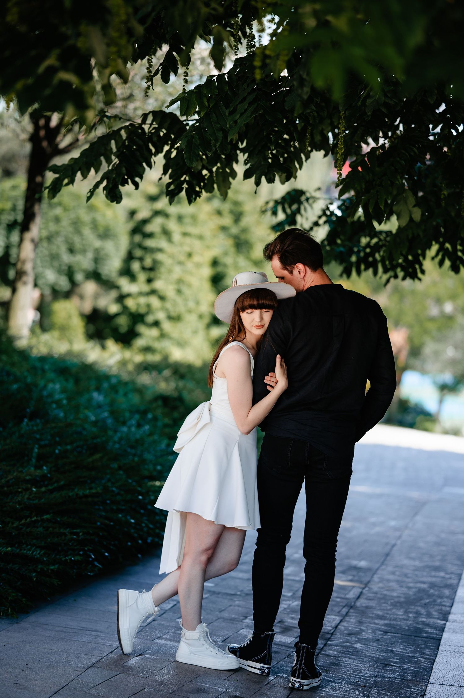 Romantic couple embracing under green tree canopy on a sunlit park pathway.