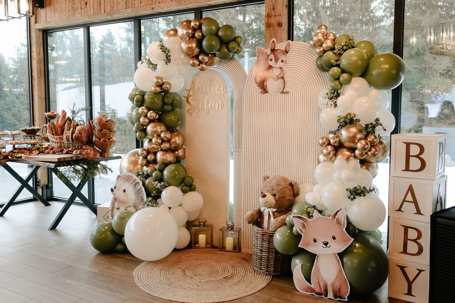 Woodland-themed baby shower backdrop with green and gold balloon arch, stuffed animals, and BABY letter blocks.