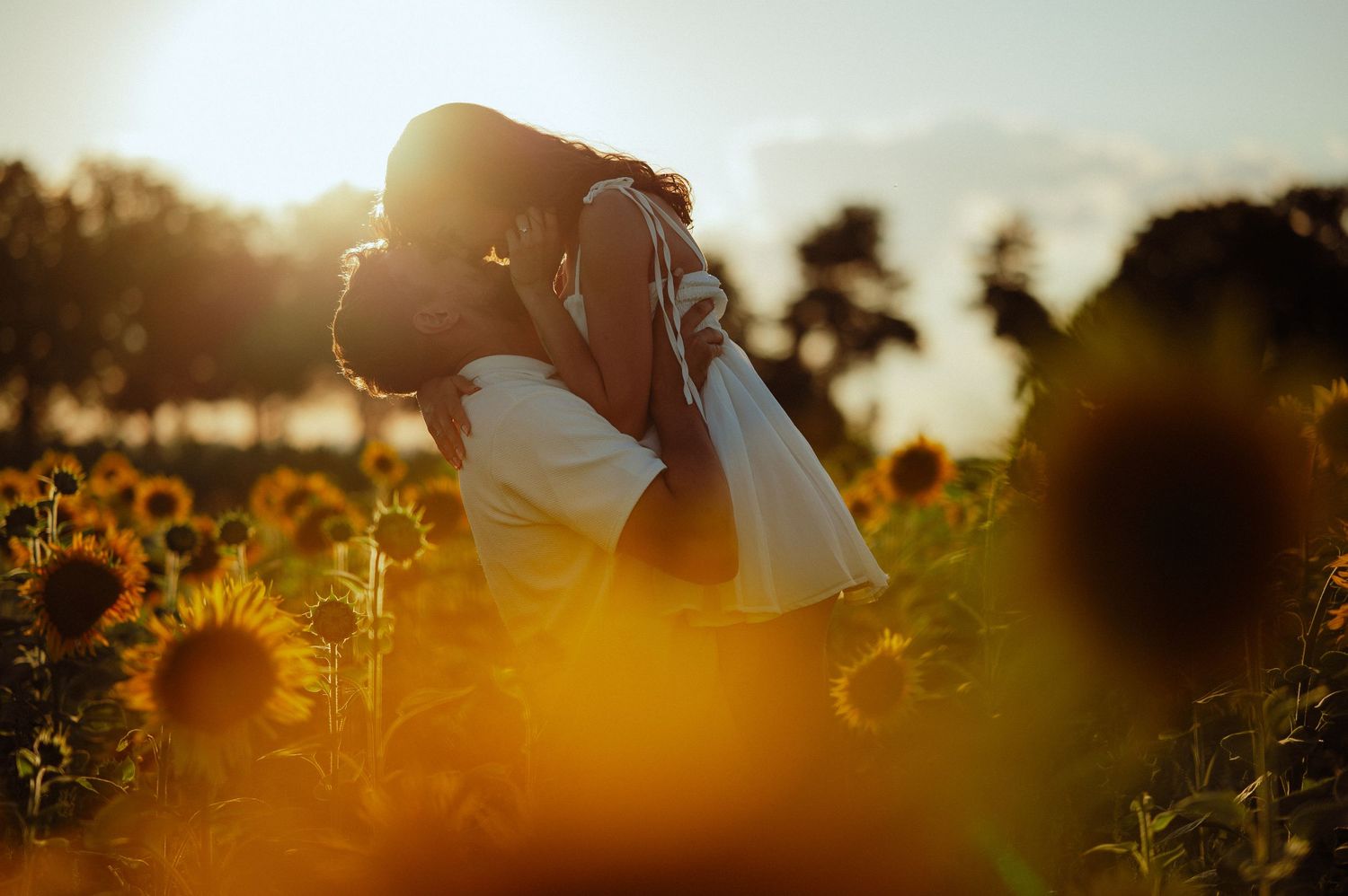 Couple sharing a romantic kiss silhouetted by golden sunset light in a sunflower field.