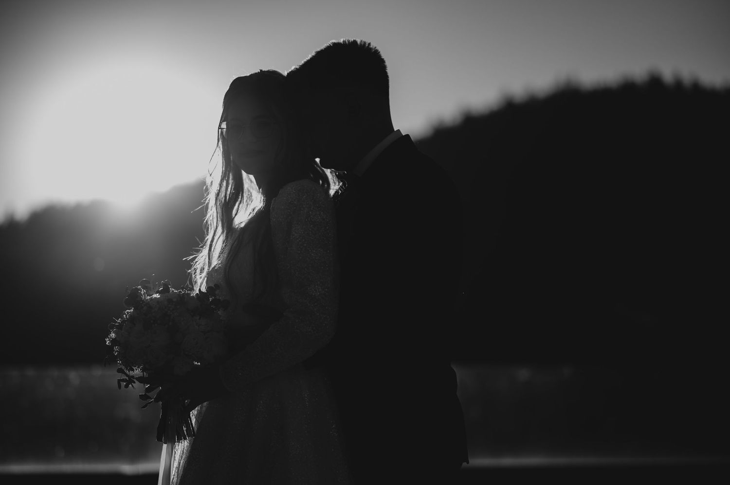 Romantic black and white silhouette of bride and groom embracing at sunset with mountains behind.