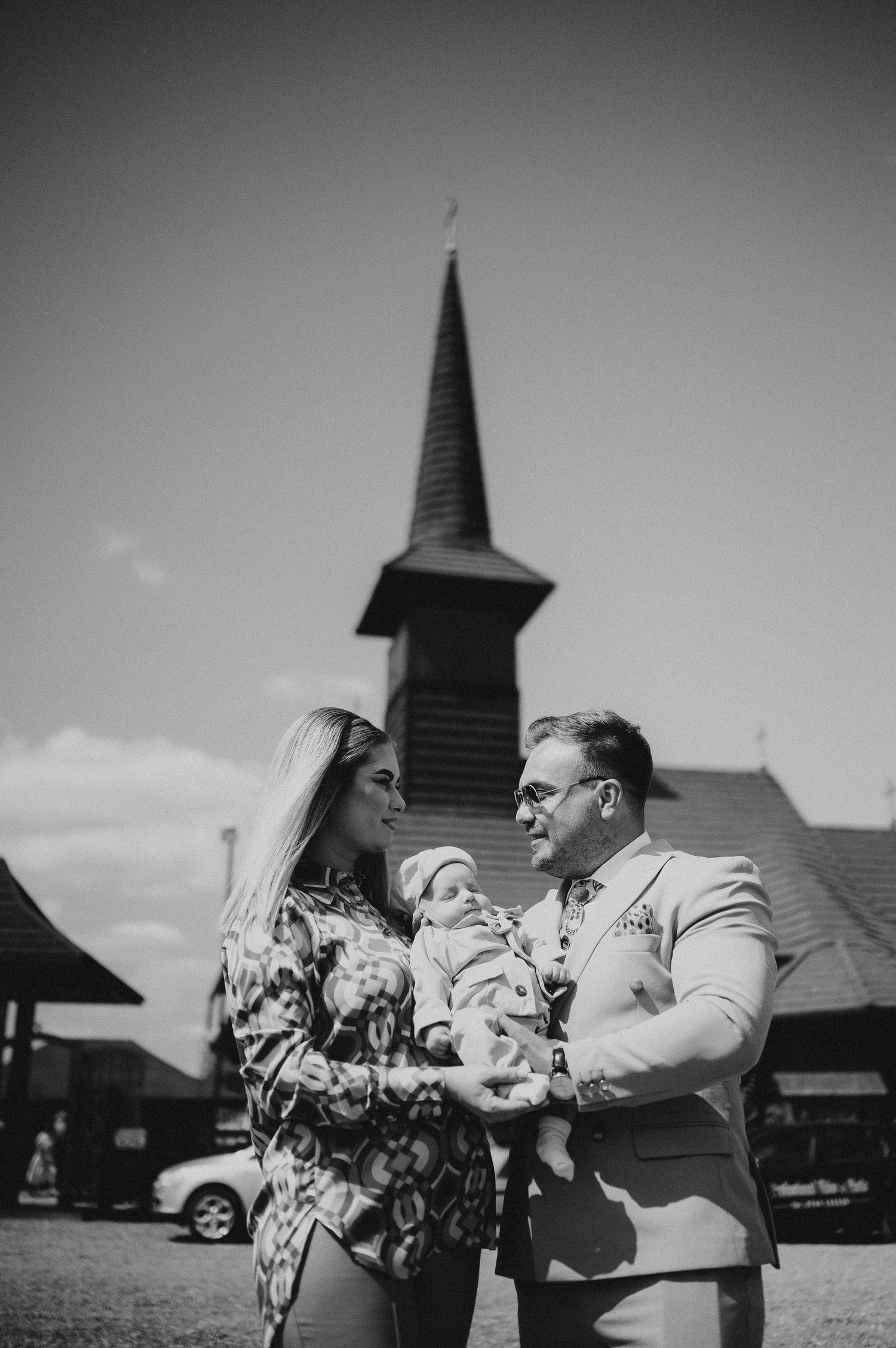 Family with baby posing outdoors in front of traditional wooden church steeple, black and white.