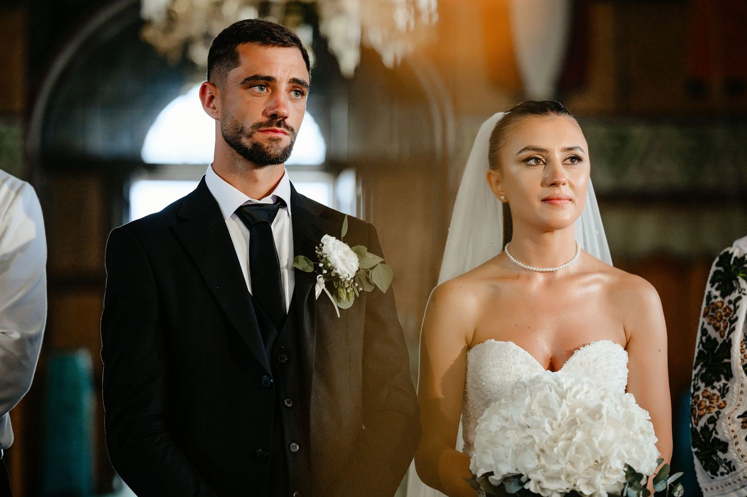 Bride and groom stand together during Orthodox church wedding ceremony, bride holding white floral bouquet.