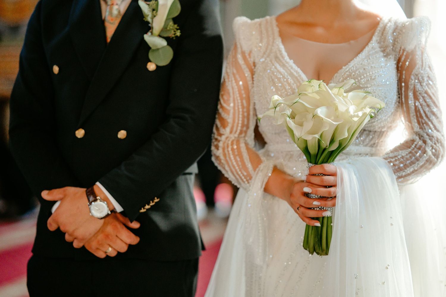 Close-up of bride and groom hands during wedding ceremony, bride holding elegant white calla lily bridal bouquet.