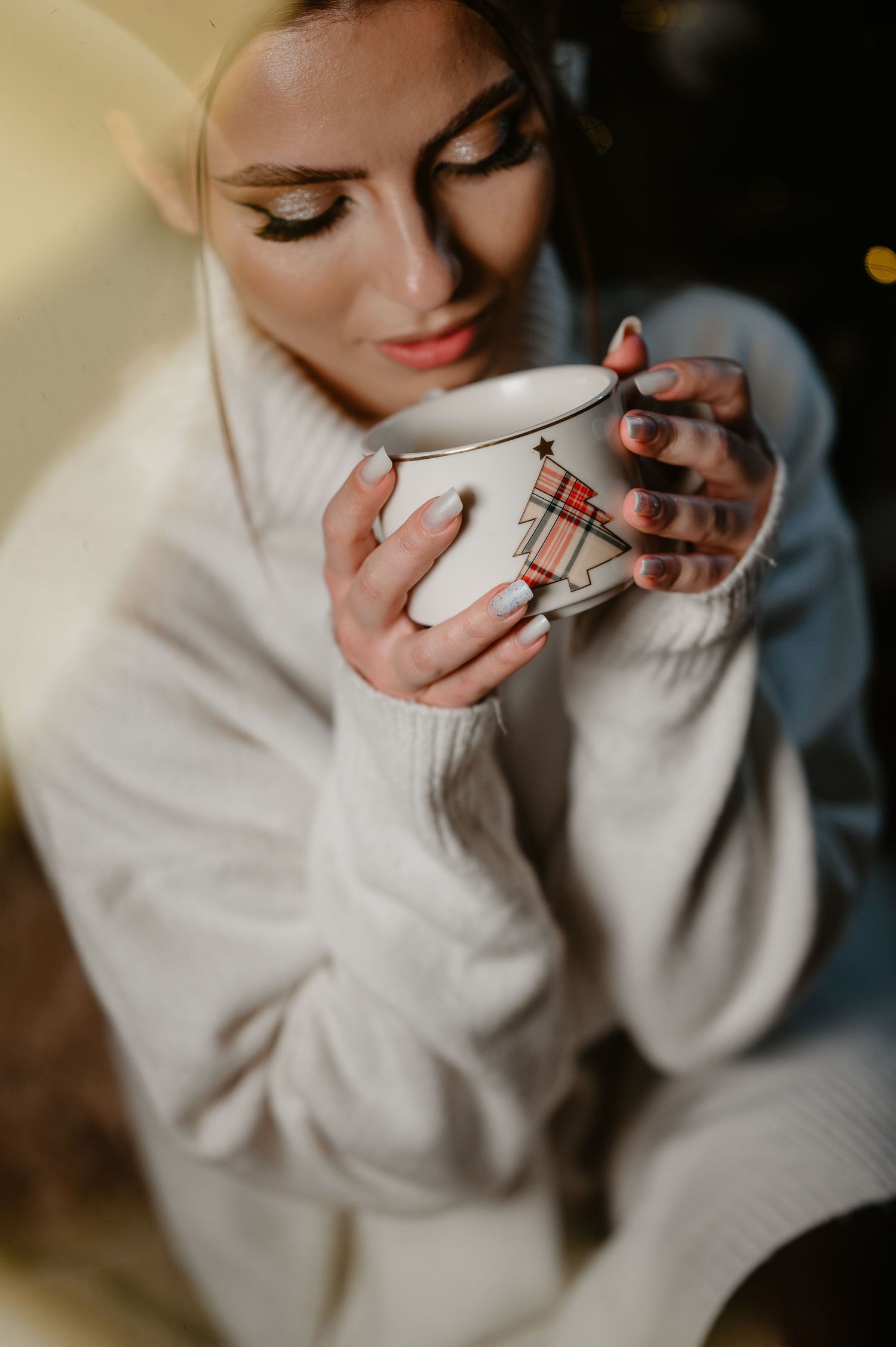 Close-up of woman in white sweater holding festive holiday mug with bokeh light.