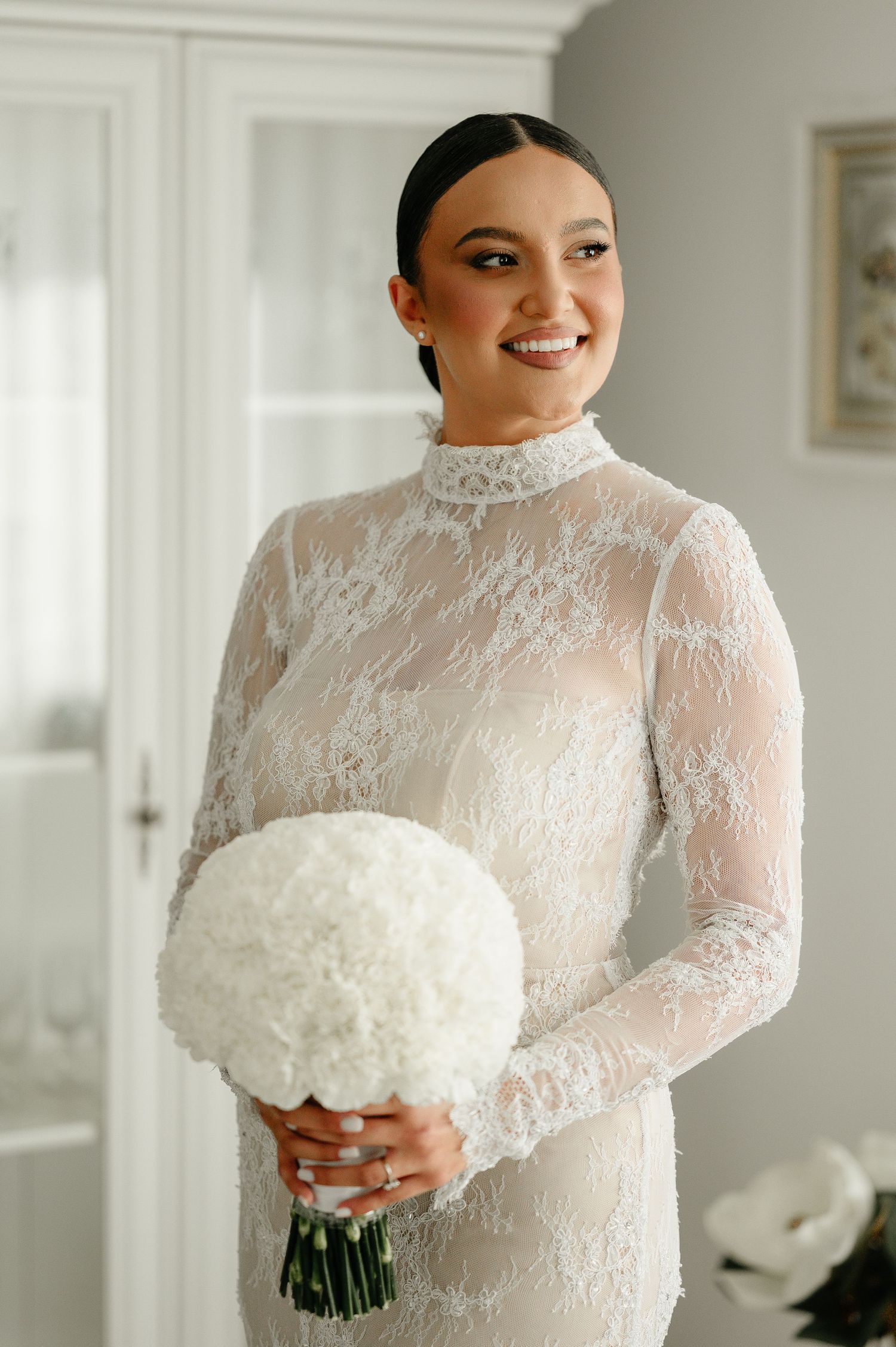 Radiant bride in high neck lace wedding dress holding round white hydrangea bouquet.