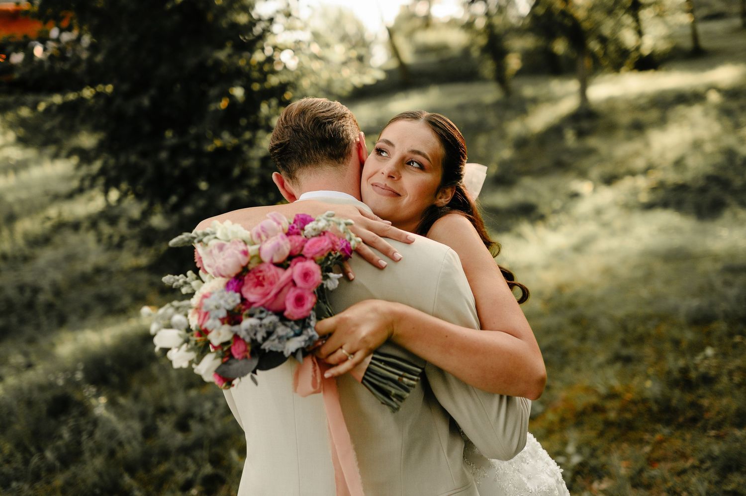 Bride embraces groom while holding a pink and white bouquet, smiling joyfully in a lush green setting.