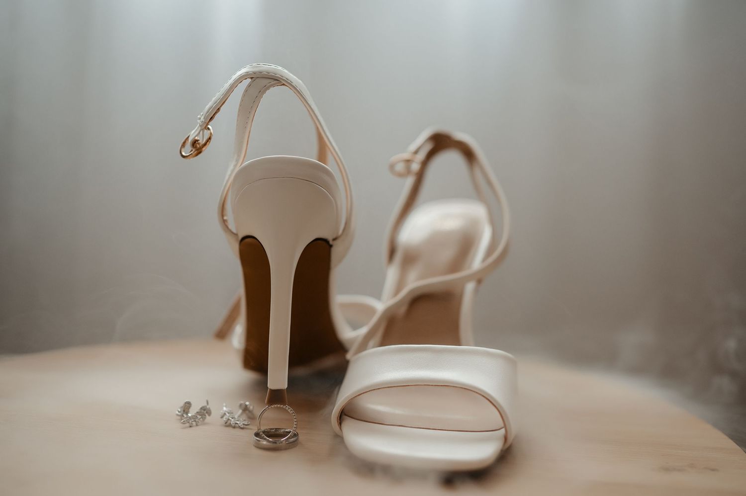 White bridal high heel sandals with ankle straps displayed alongside wedding rings on a neutral surface.