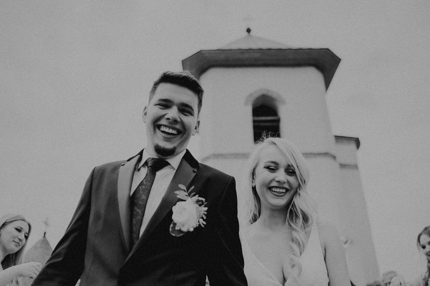 Black and white photo of laughing newlyweds exiting church with a bell tower in the background.