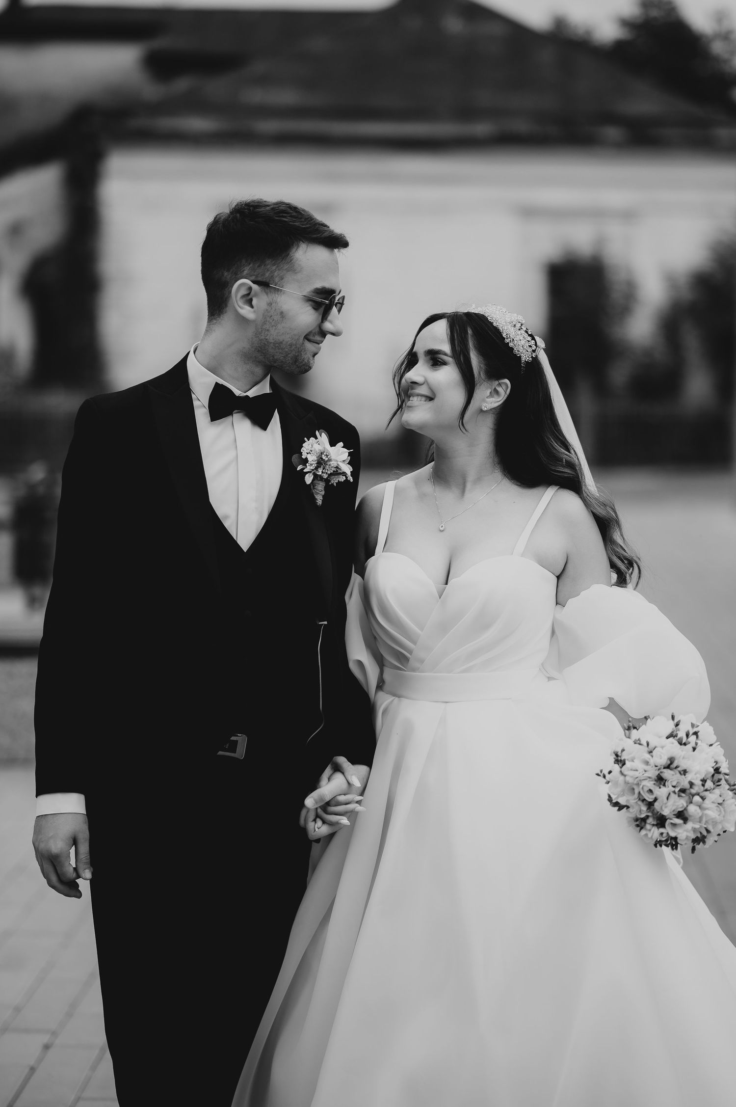 Black and white wedding photo of smiling bride in gown holding bouquet and groom in tuxedo holding hands outdoors.