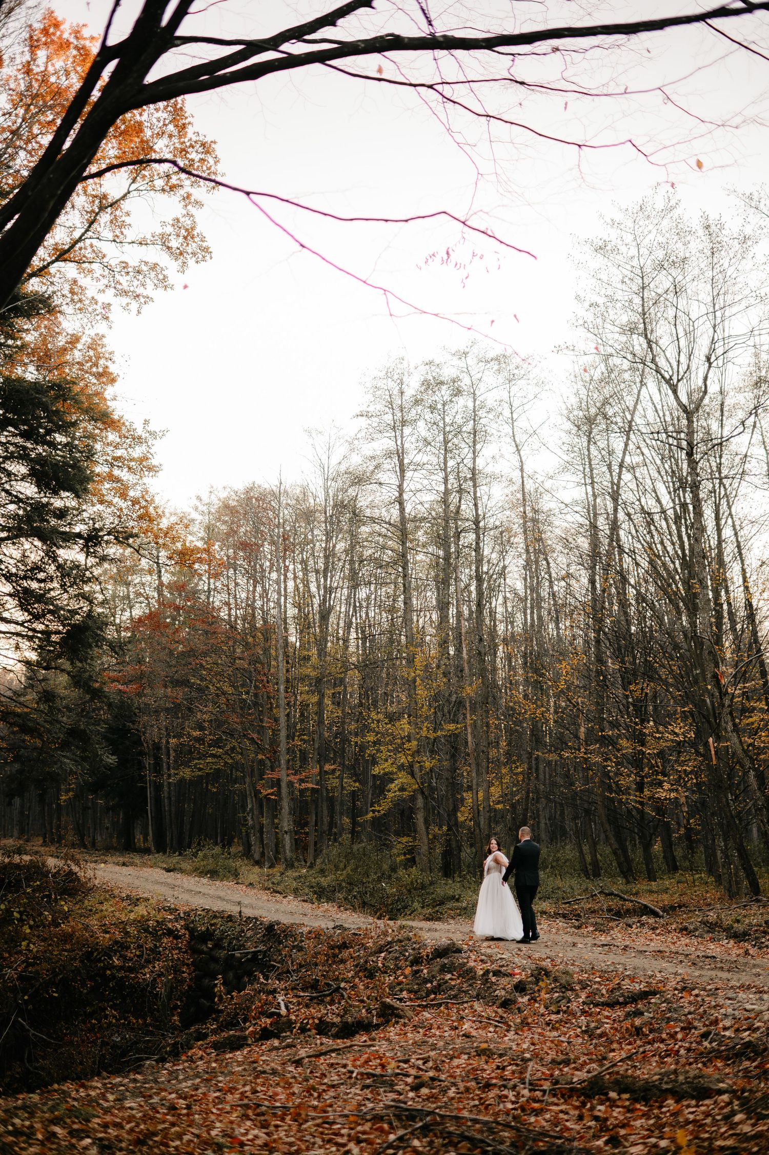 Bride and groom embracing on a forest path surrounded by autumn foliage and tall bare trees on their wedding day.
