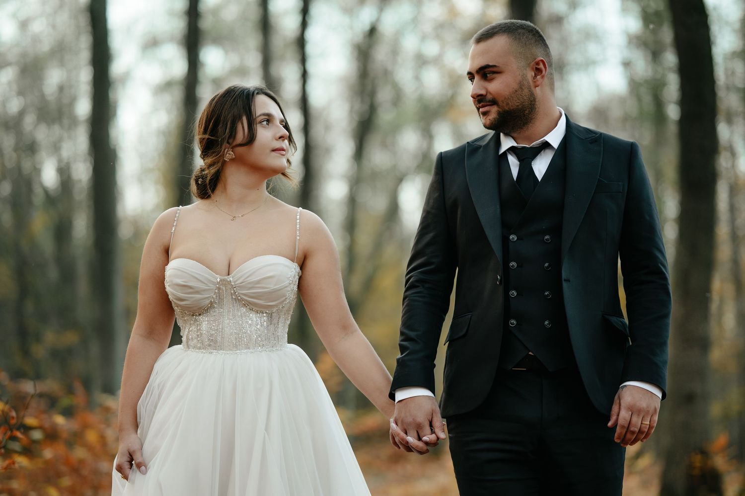 Bride and groom holding hands walking through autumn forest, she in white gown, he in black tuxedo.