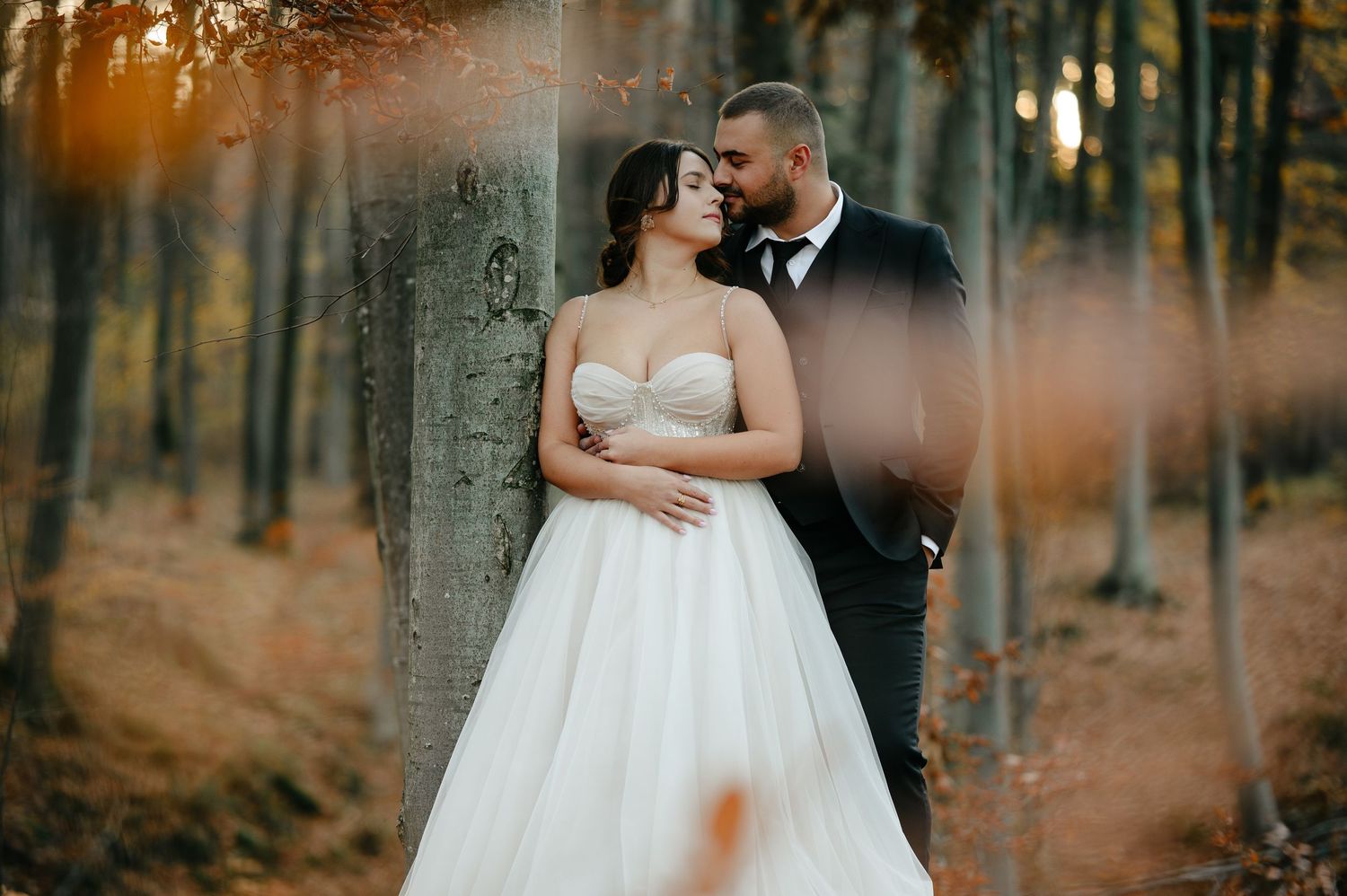 Romantic couple in wedding attire embracing in an autumn forest, bride in white gown and groom in black suit.