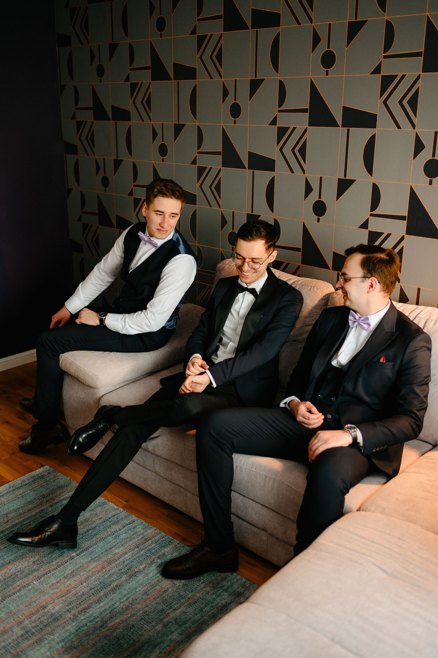Three groomsmen in formal attire sitting on a couch, laughing and chatting before a wedding ceremony.