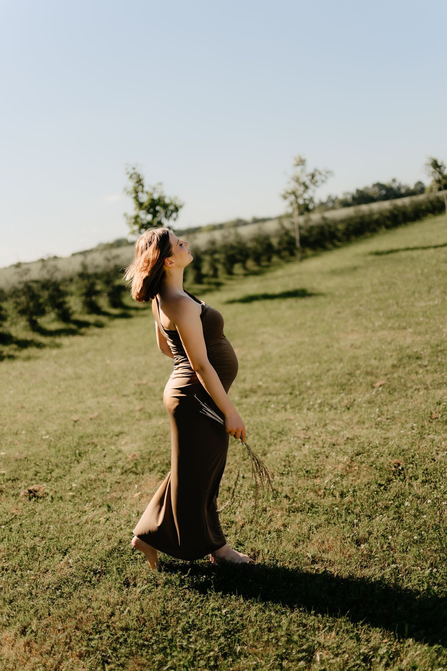 Pregnant woman in dark maxi dress walking on green hillside in sunny outdoor maternity photo.