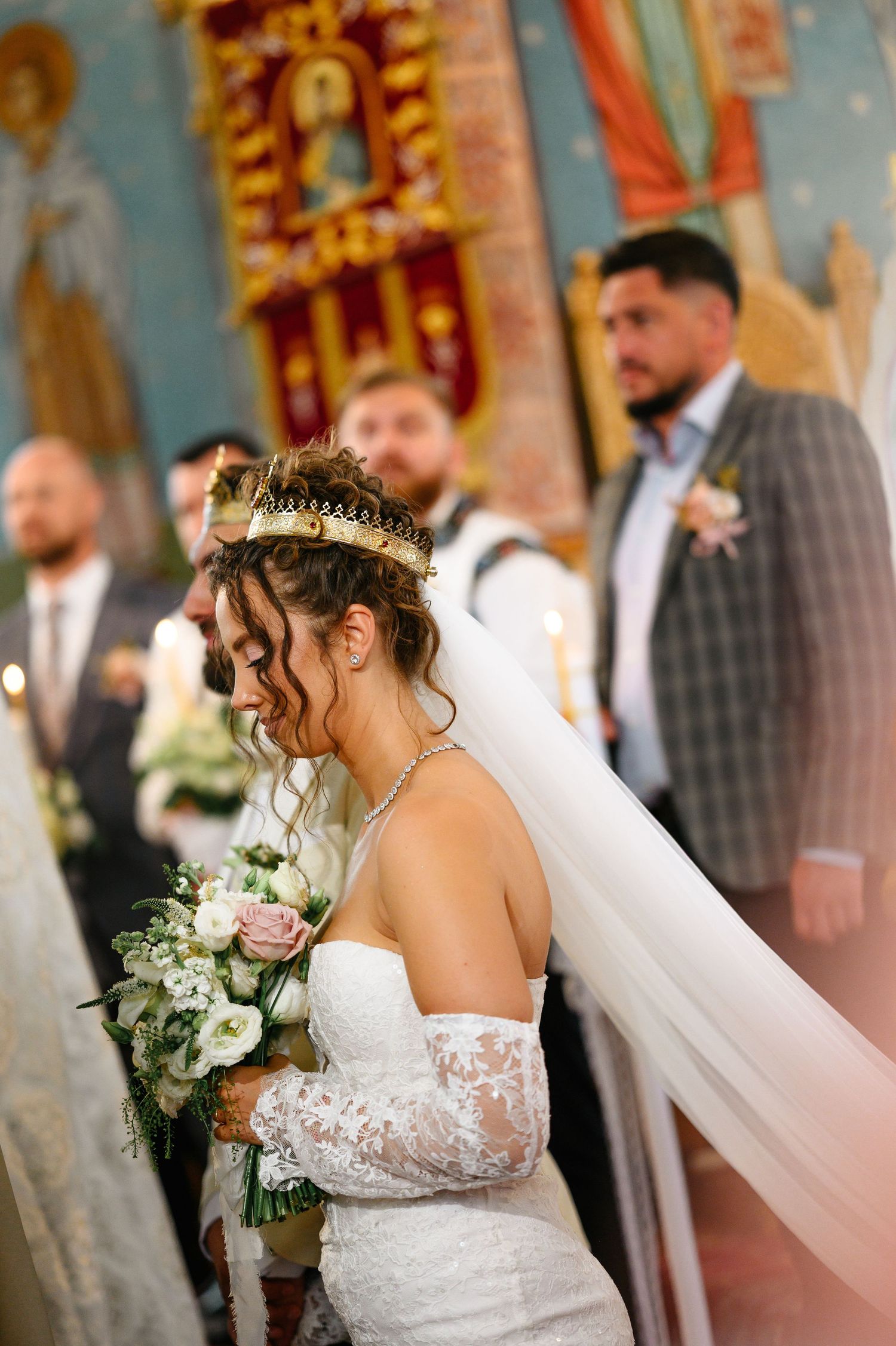 Bride in lace wedding dress and crown veil holds bouquet during Orthodox church ceremony.
