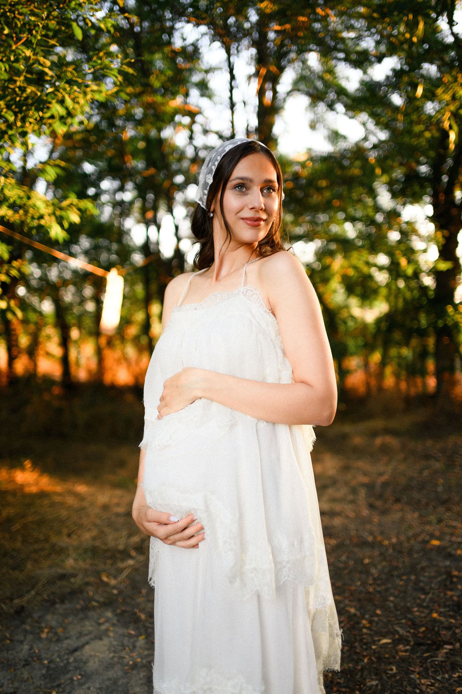 Pregnant woman in white lace dress and headband cradling her baby bump outdoors in a sunlit forest.