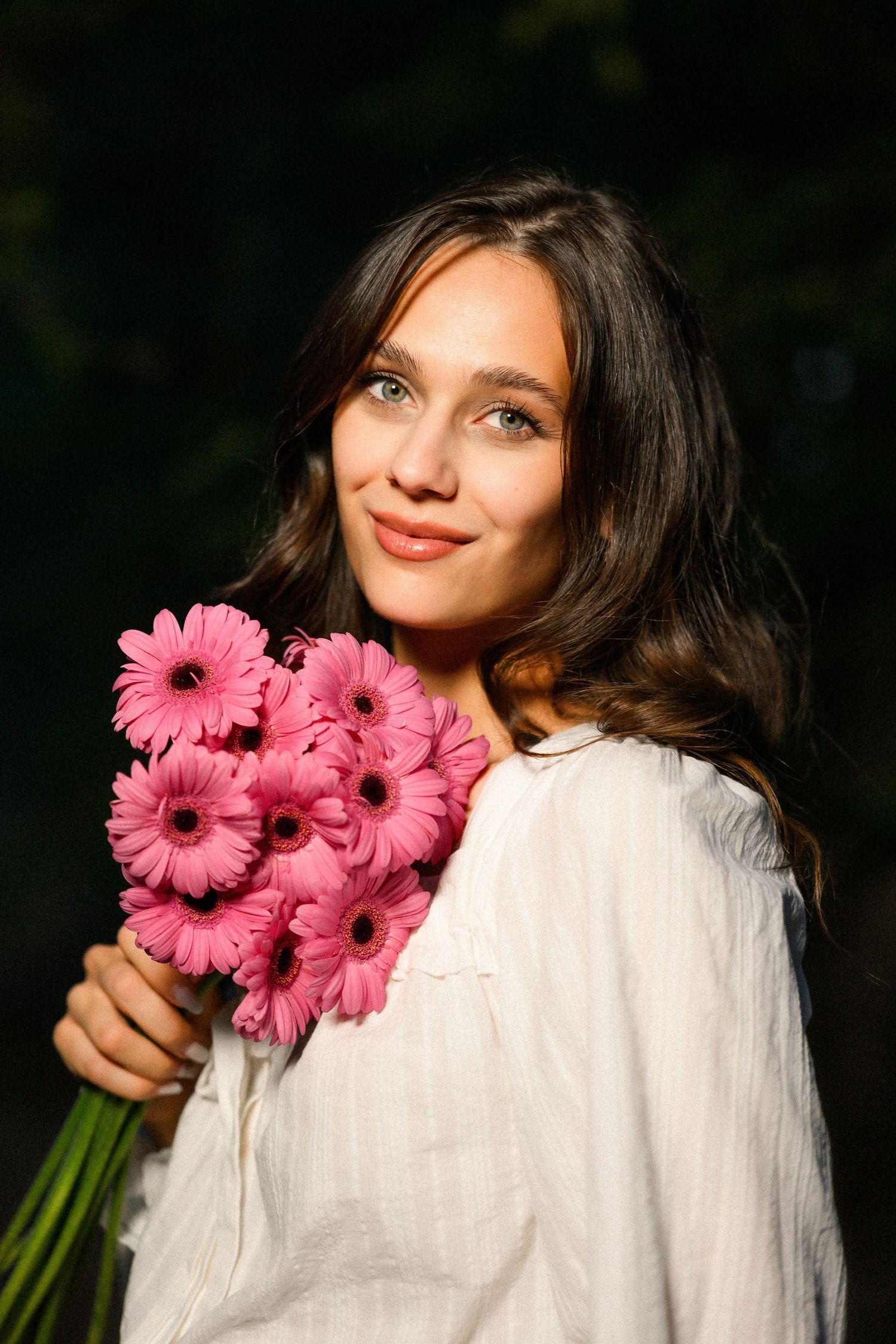 Young woman with dark wavy hair holding a bouquet of pink gerbera daisies, wearing a white blouse outdoors.