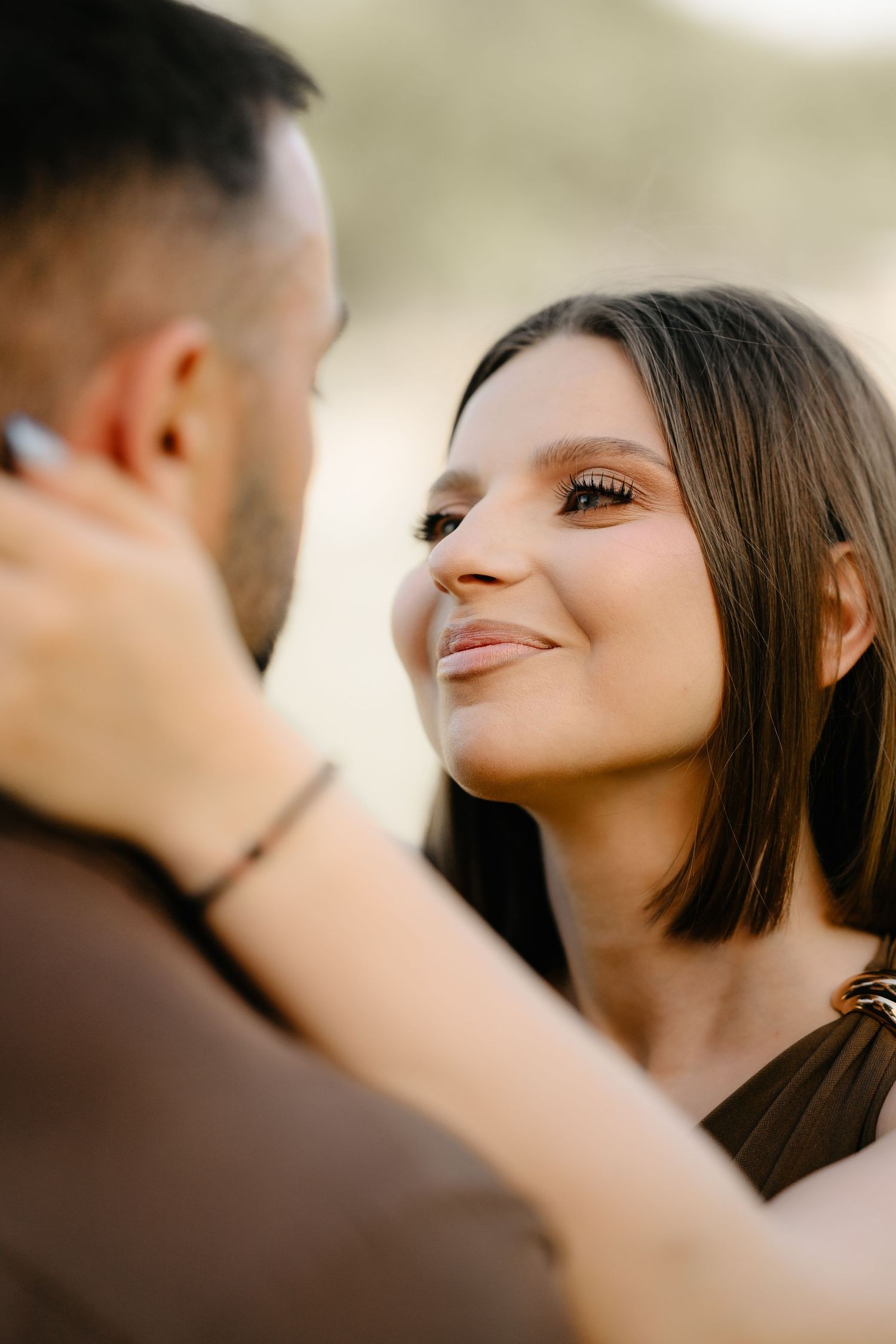 Smiling brunette woman gazes lovingly at man while embracing him outdoors in warm natural light.