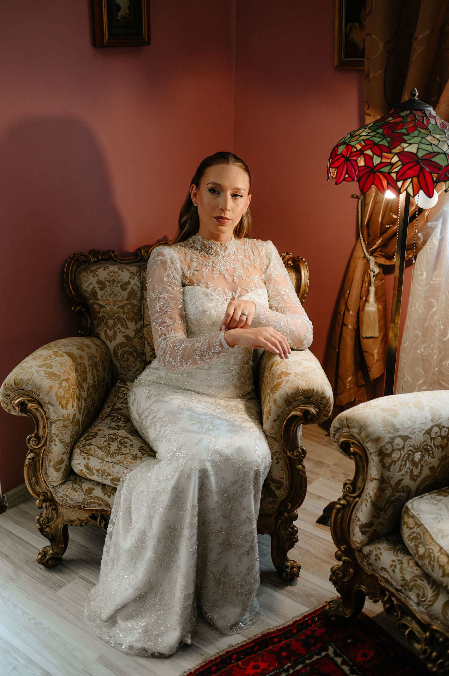 Bride in long-sleeve lace wedding gown seated on ornate gold armchair in elegant vintage room.