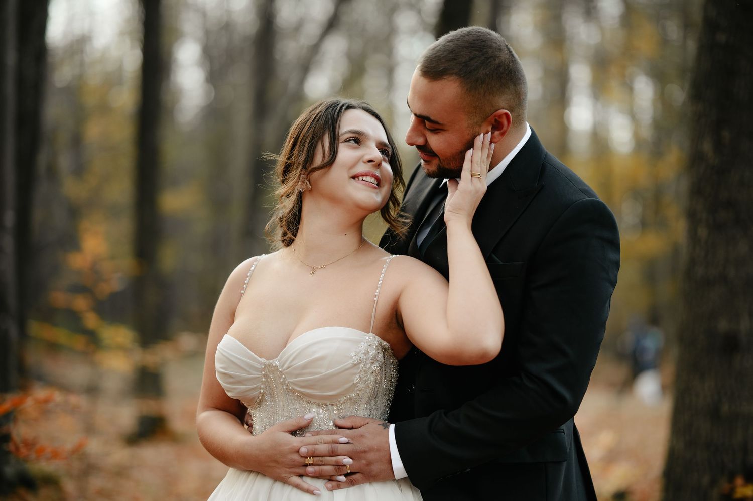 Bride and groom share a romantic moment in autumn forest, she in strapless gown, he in black tuxedo.