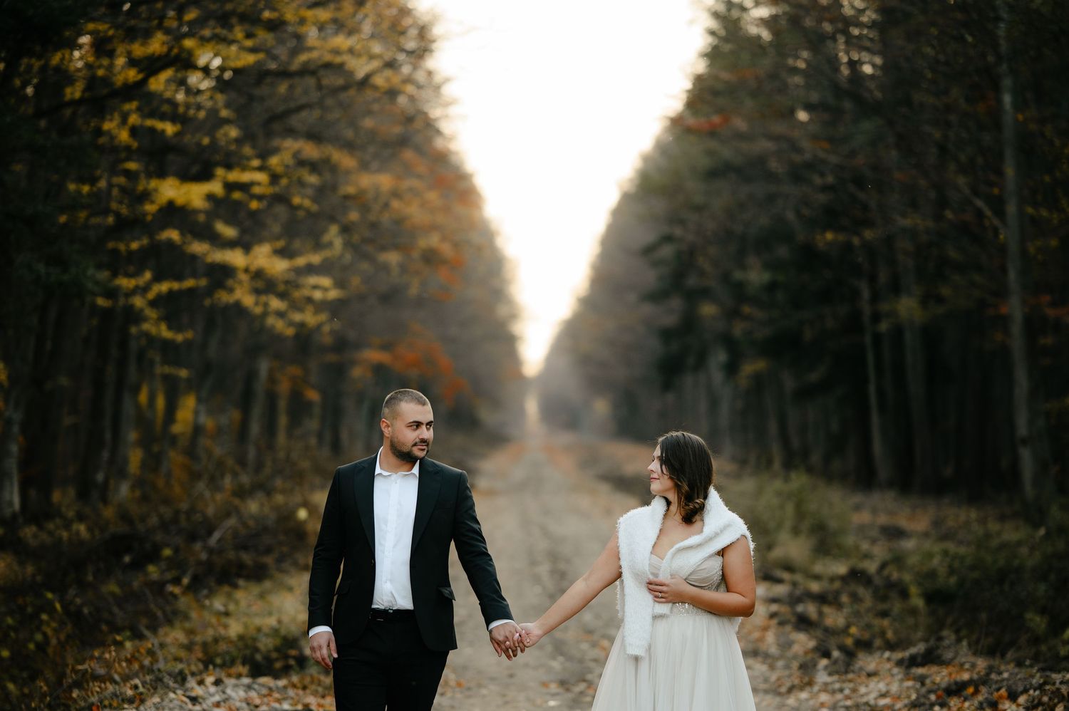 Couple in wedding attire holding hands on a forest path surrounded by autumn trees.