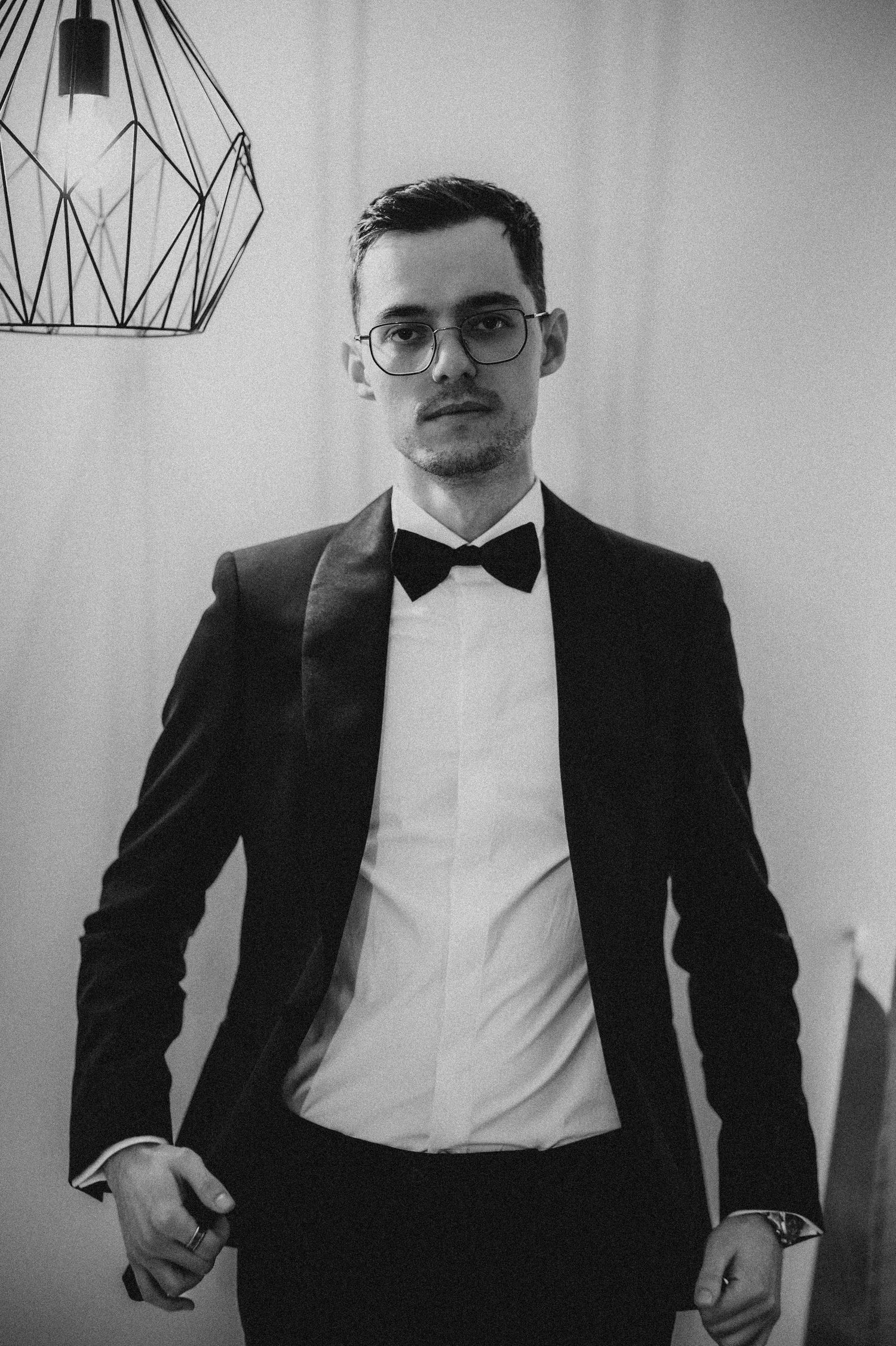 Young man in black tuxedo with bow tie and glasses posing confidently in a black and white portrait photo.