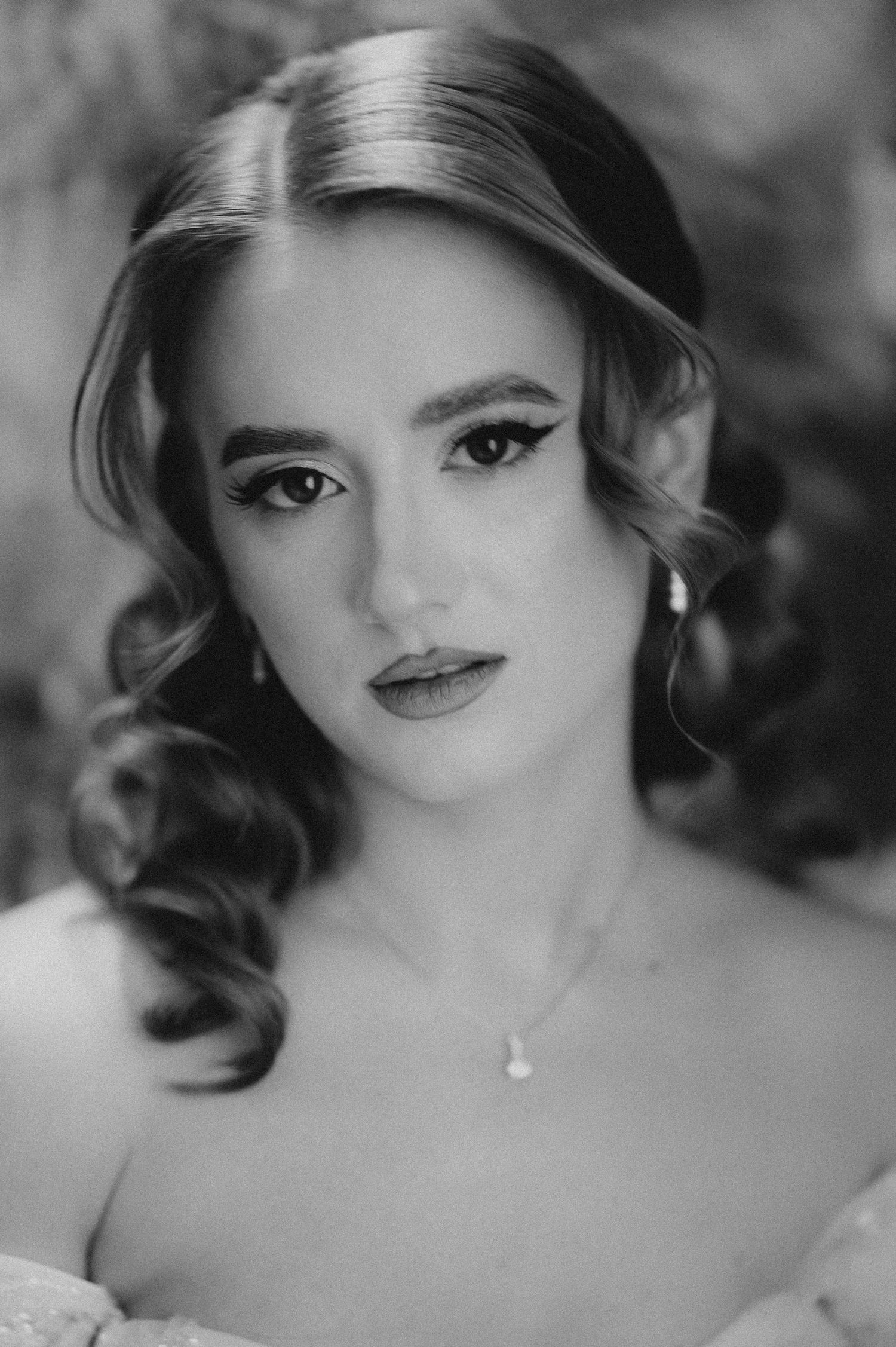 Black and white bridal portrait of a young woman with vintage curls, dramatic eye makeup, and a delicate pearl necklace.