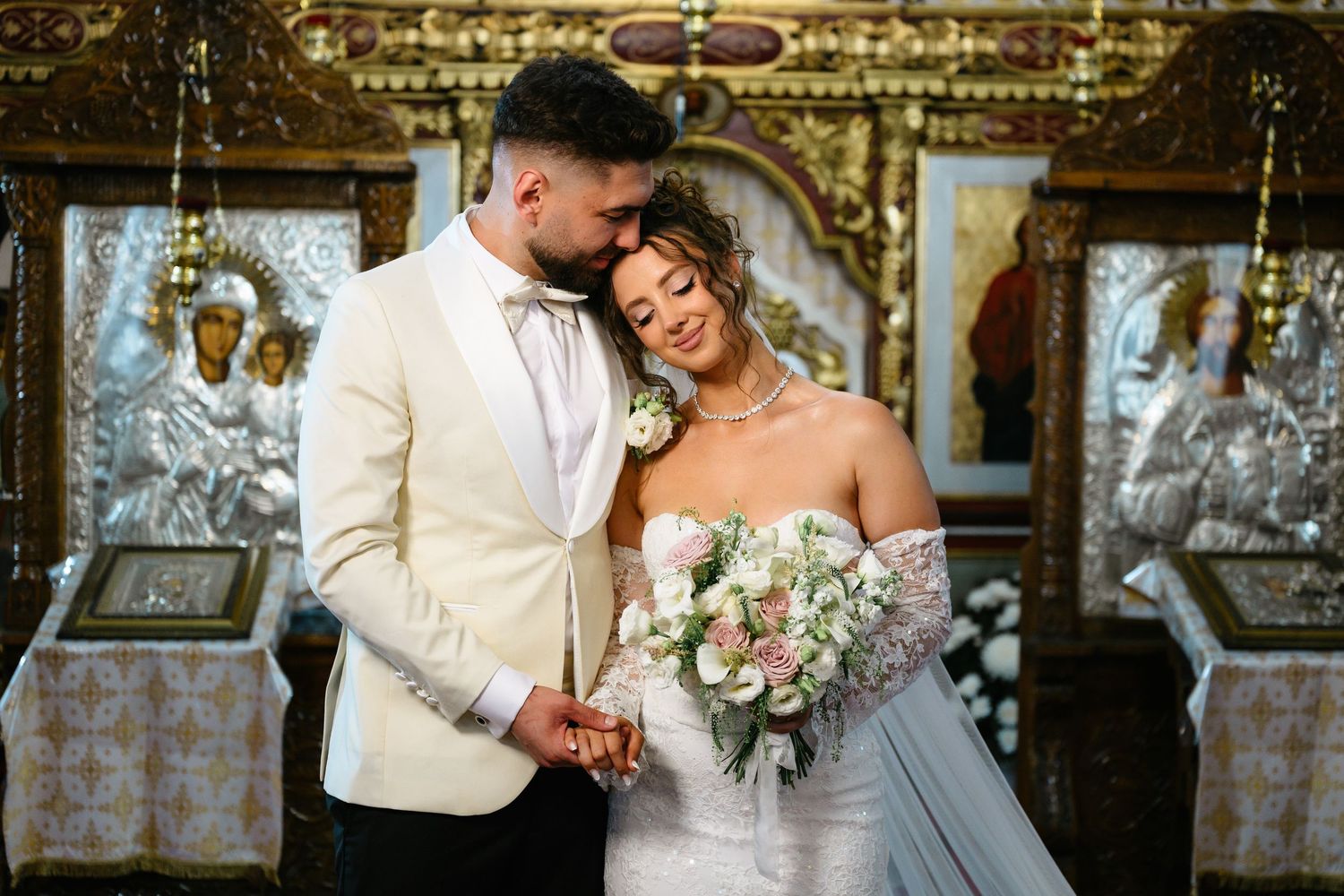 Bride and groom embrace tenderly in an Orthodox church, bride holding pink bouquet, groom in ivory tuxedo.