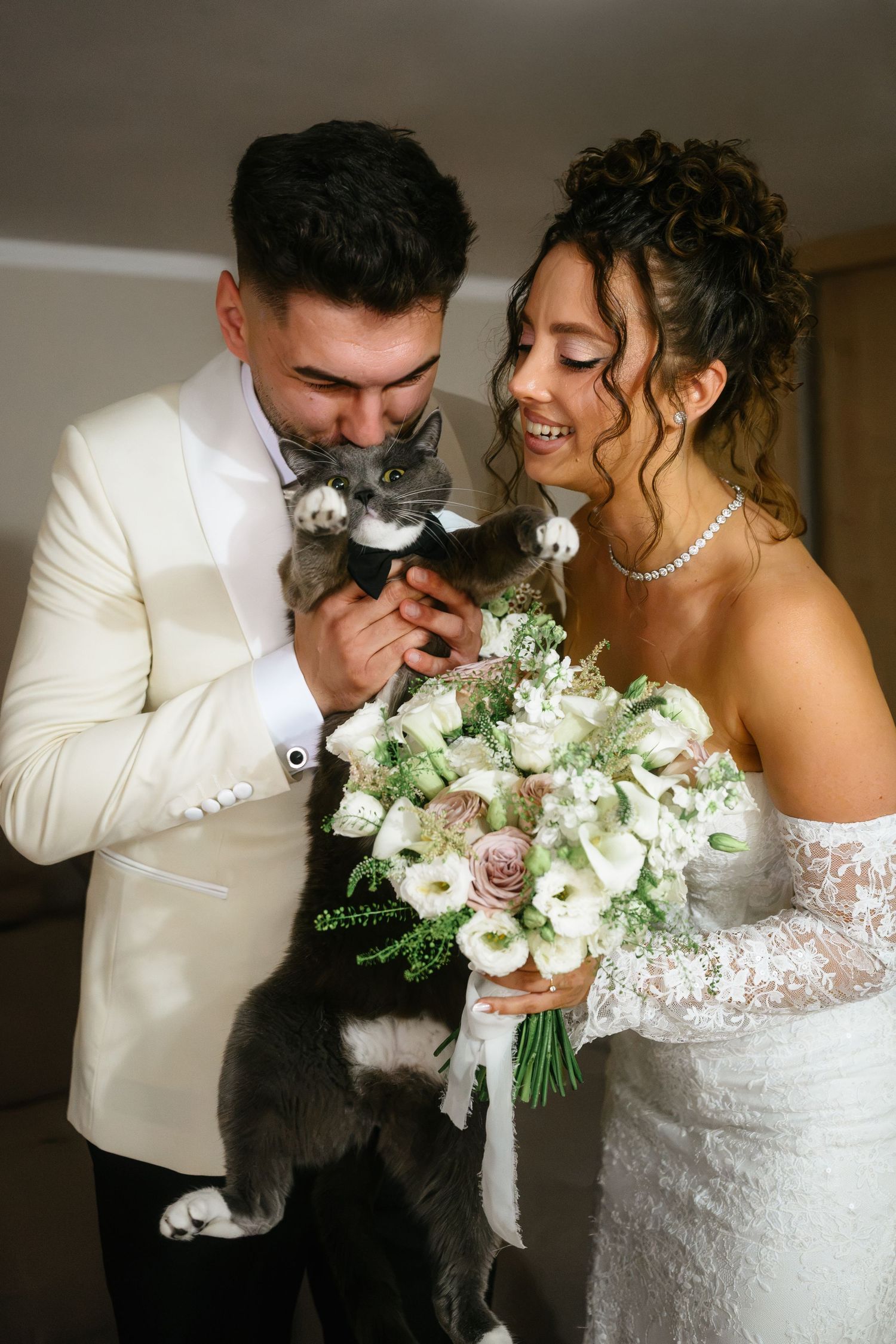 Newlyweds in wedding attire cuddling a gray cat while holding a white rose bouquet.