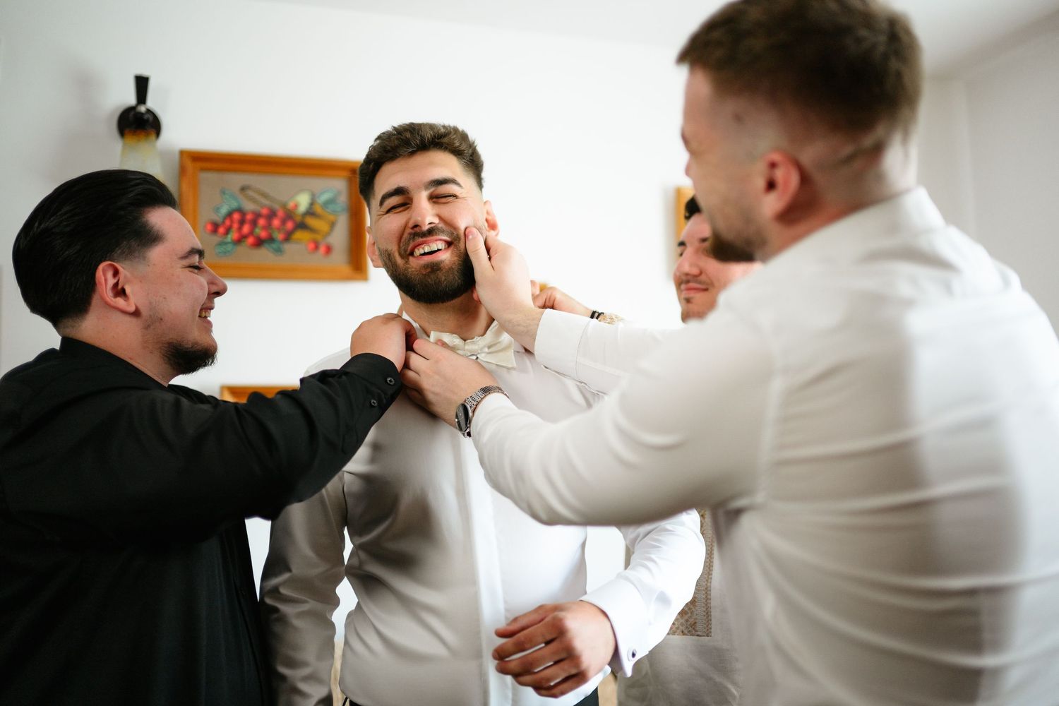 Groomsmen helping groom get dressed, adjusting shirt and tie while smiling together before wedding ceremony.