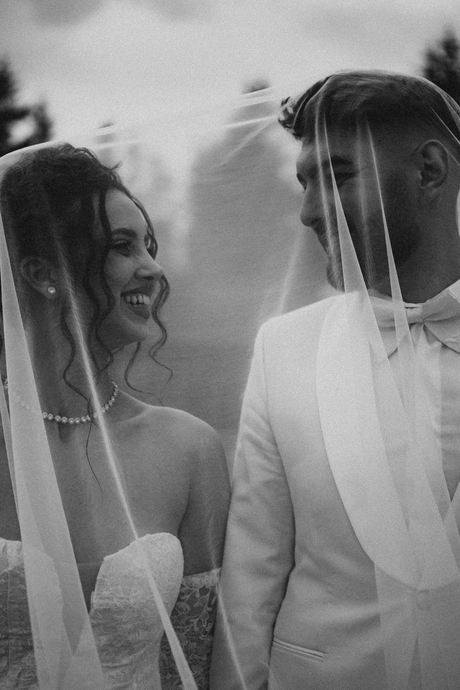 Black and white photo of smiling bride and groom under wedding veil, bride wearing pearl necklace.