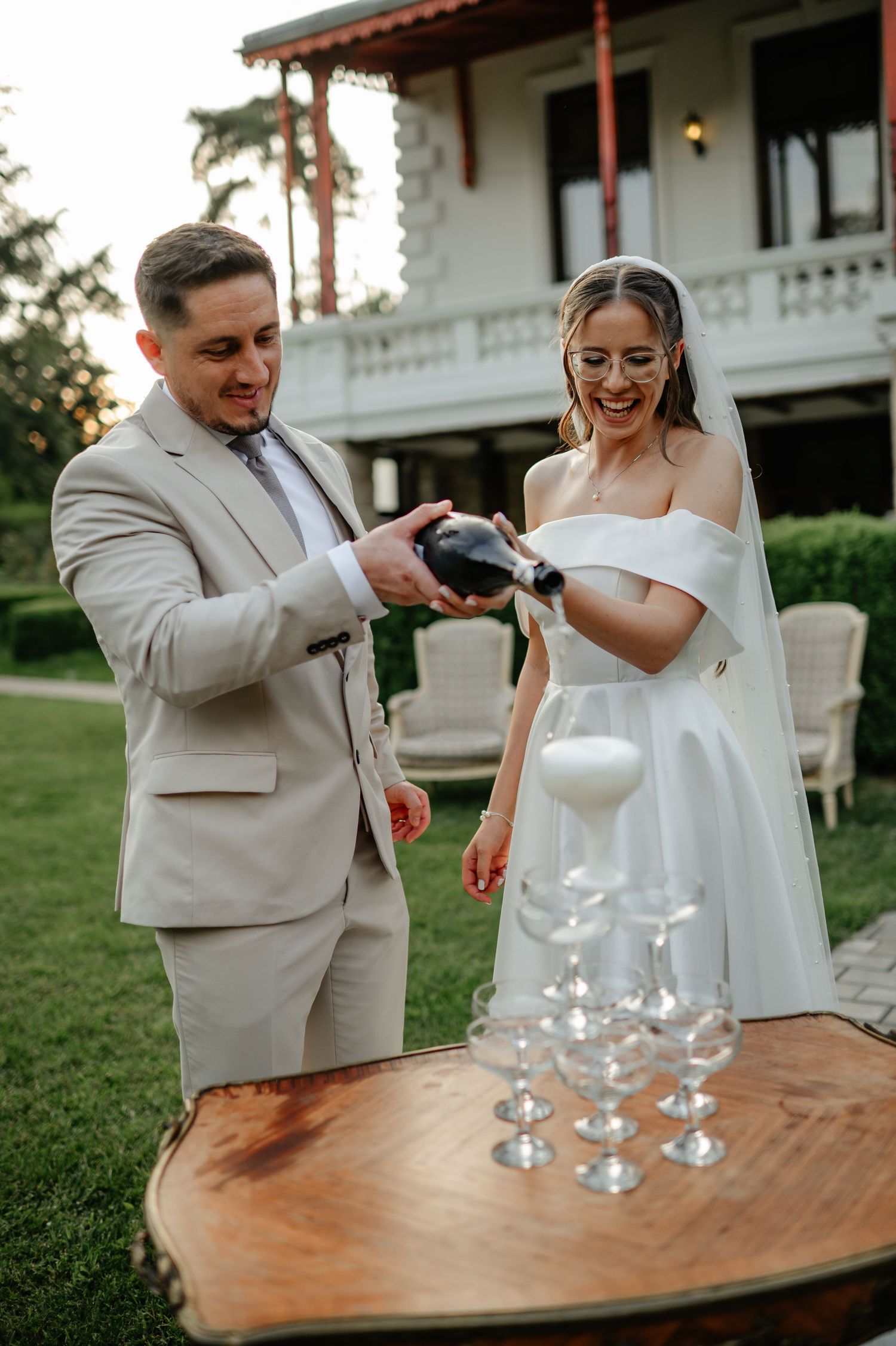 Groom in beige suit pouring champagne tower with laughing bride in off-shoulder gown outdoors.