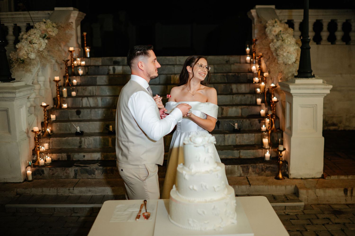 Bride and groom cut elegant white tiered wedding cake on candlelit staircase at night reception.