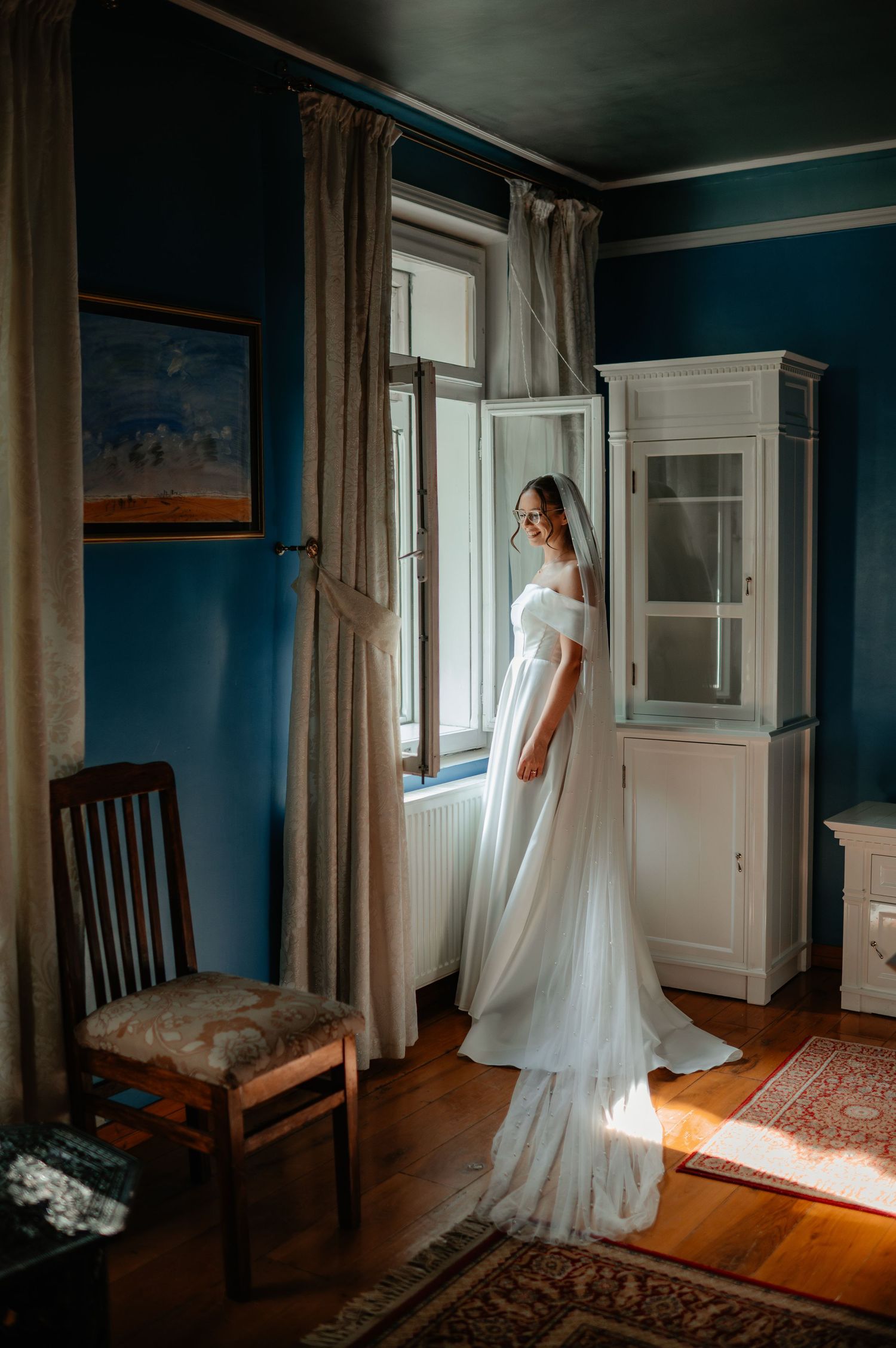 Bride in off-shoulder white gown with veil standing by window in blue room.