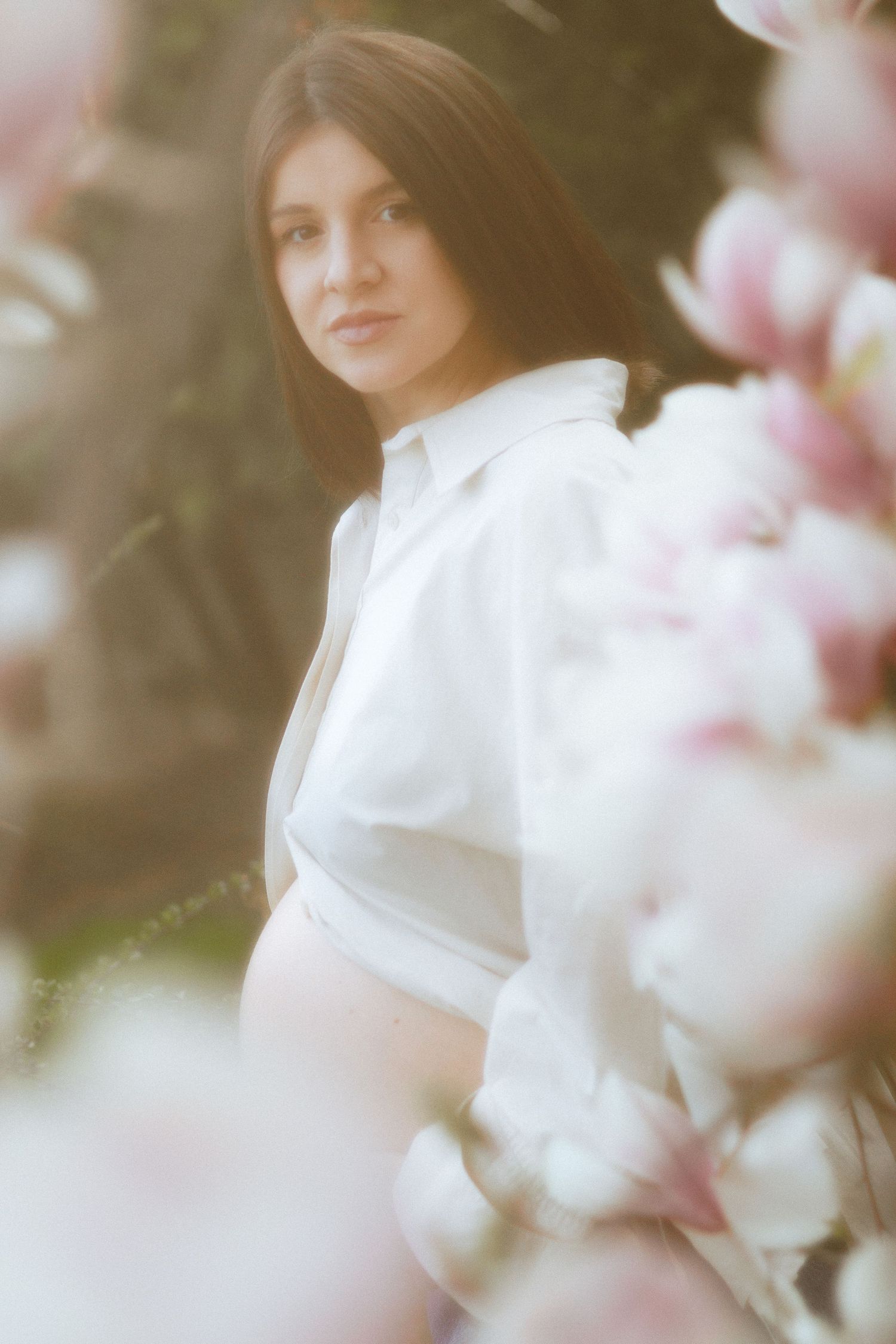 Pregnant woman in white shirt surrounded by pink magnolia blossoms in soft dreamy light.