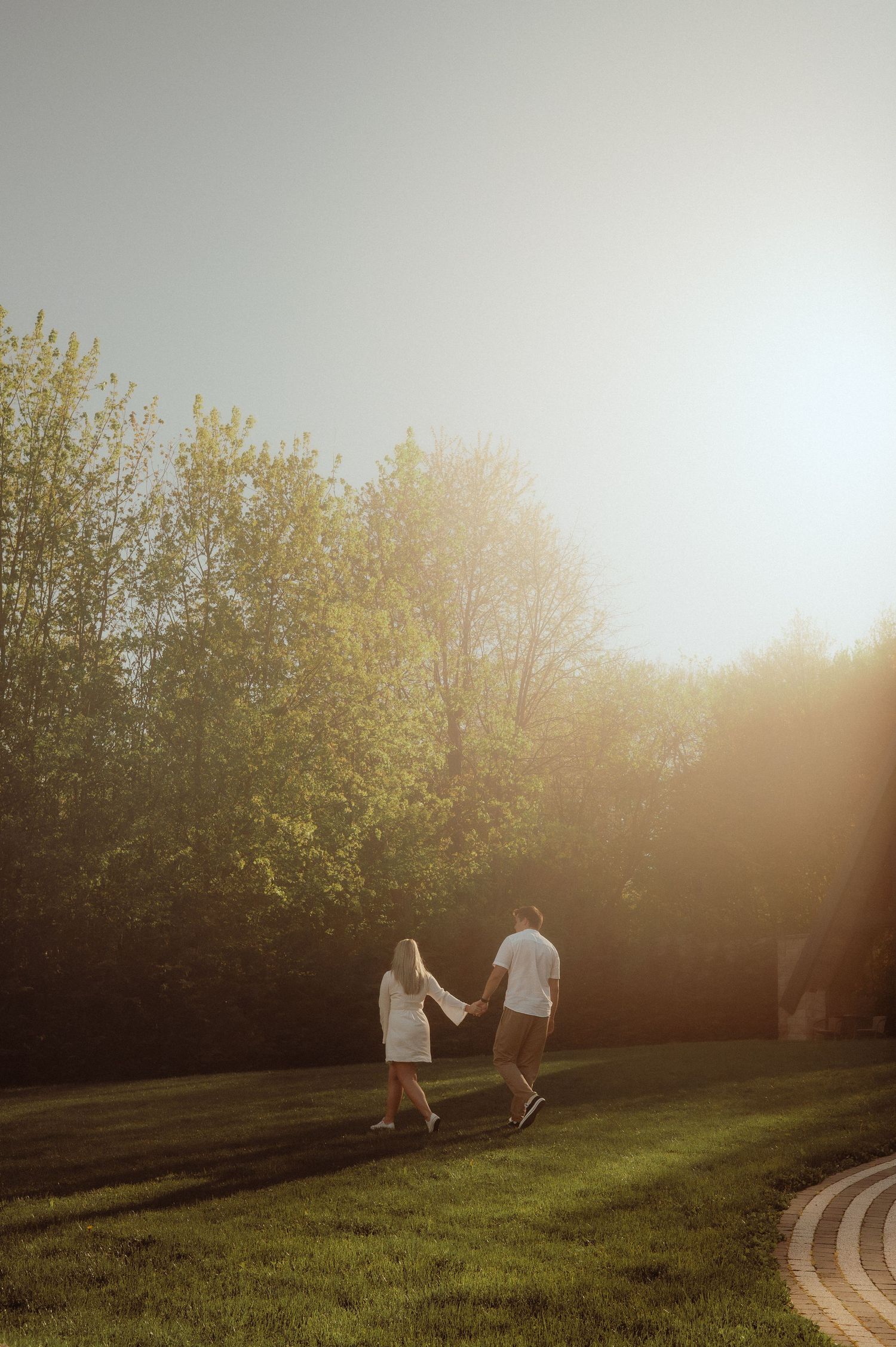 Couple strolling hand in hand across sunlit lawn with glowing golden backlight.