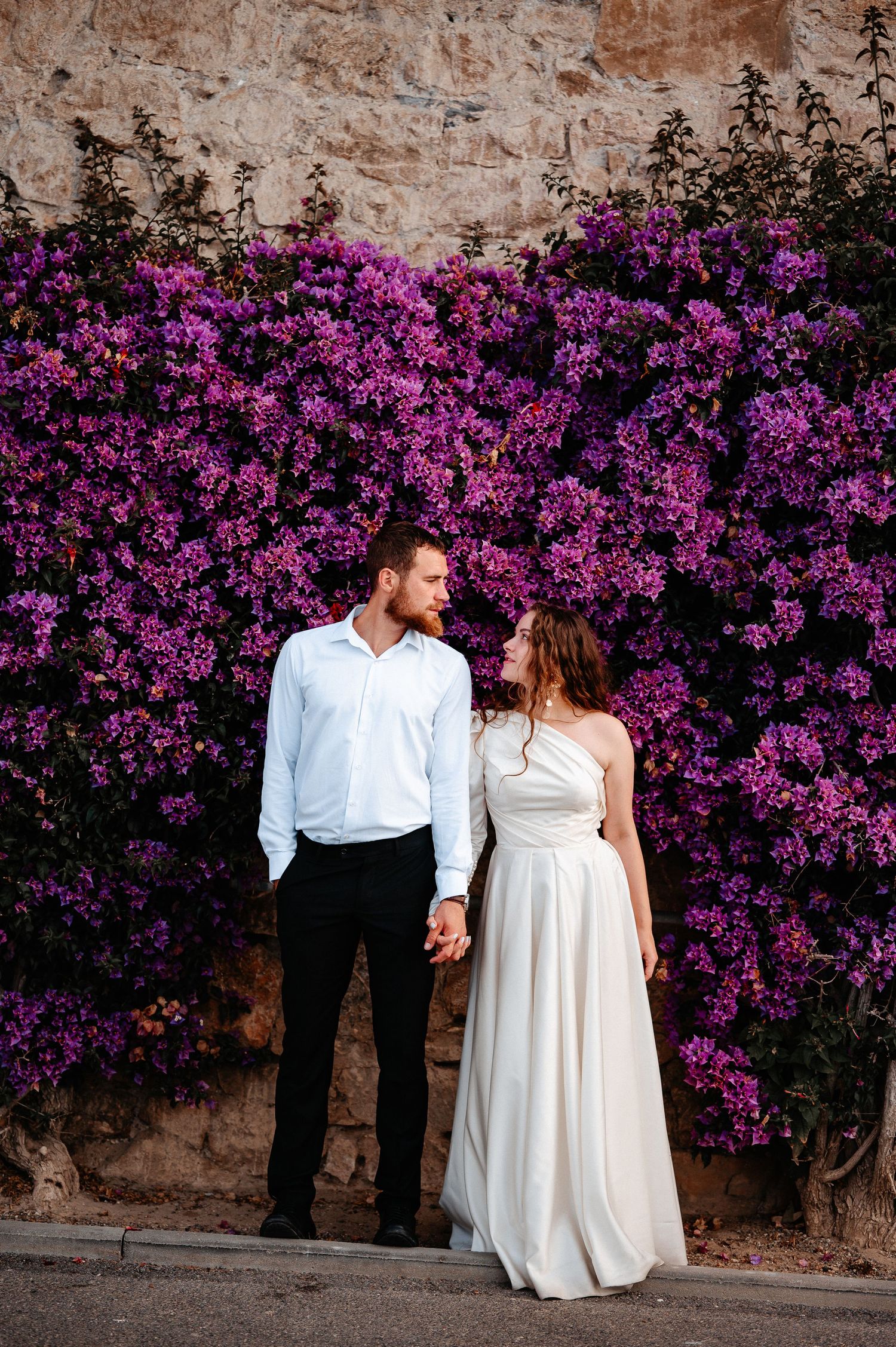 Wedding couple holds hands gazing at each other before a vibrant purple bougainvillea flower wall.