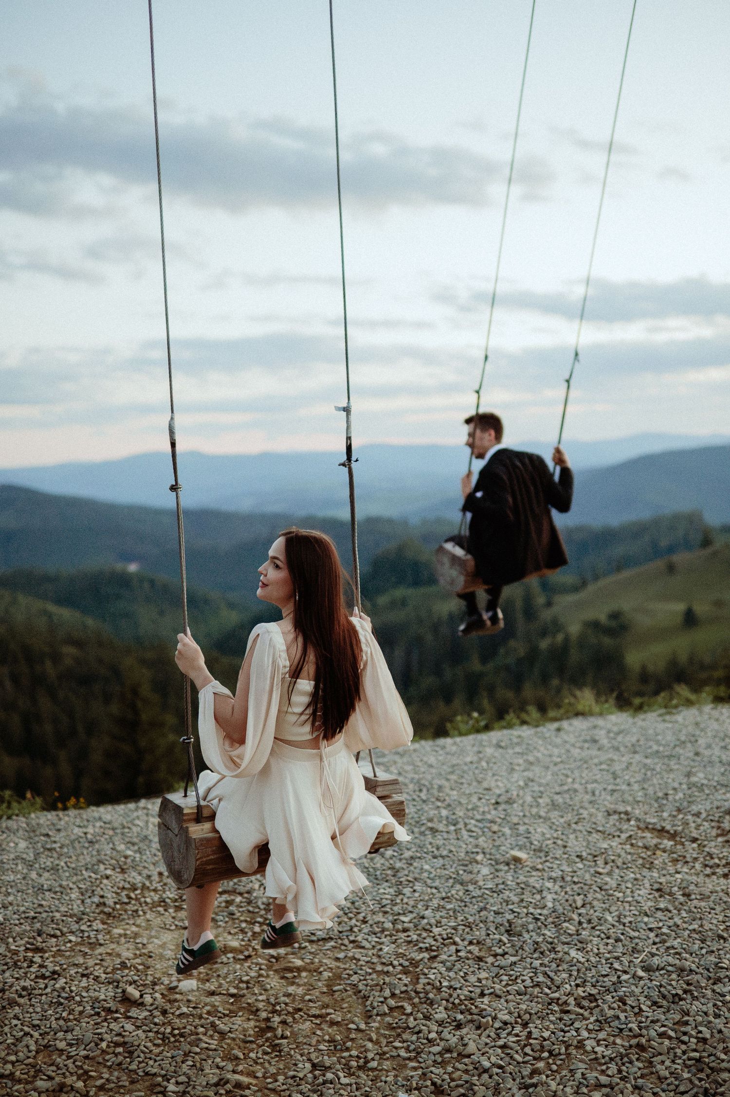 Couple sitting on wooden swings overlooking scenic mountain valley at dusk.
