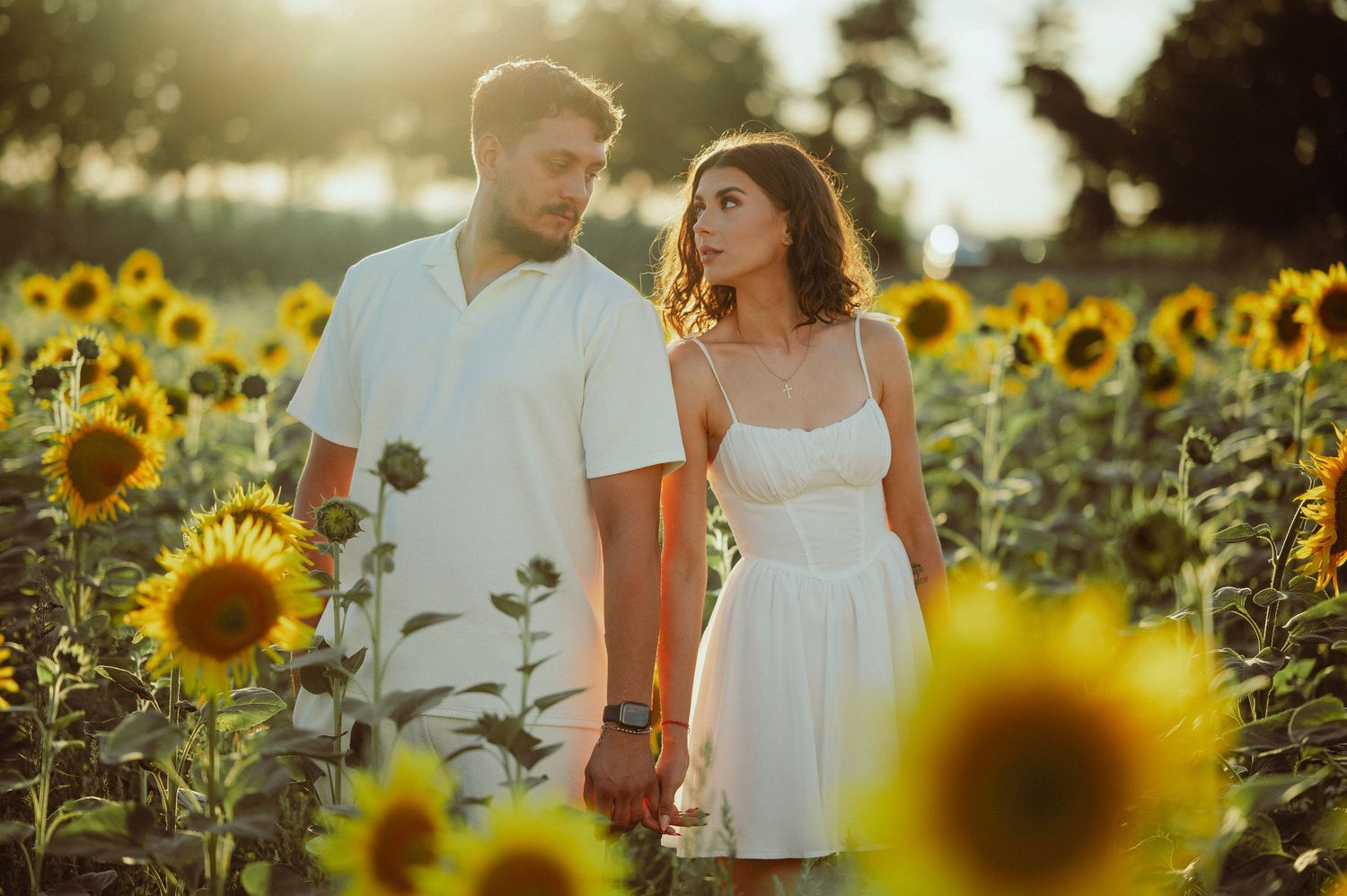 Couple holding hands and gazing at each other surrounded by blooming sunflowers at sunset.