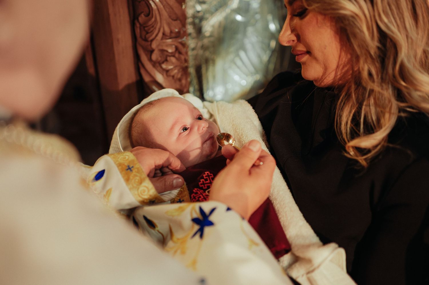 Priest holding a newborn baby wrapped in a decorative blanket during an Orthodox baptism ceremony.