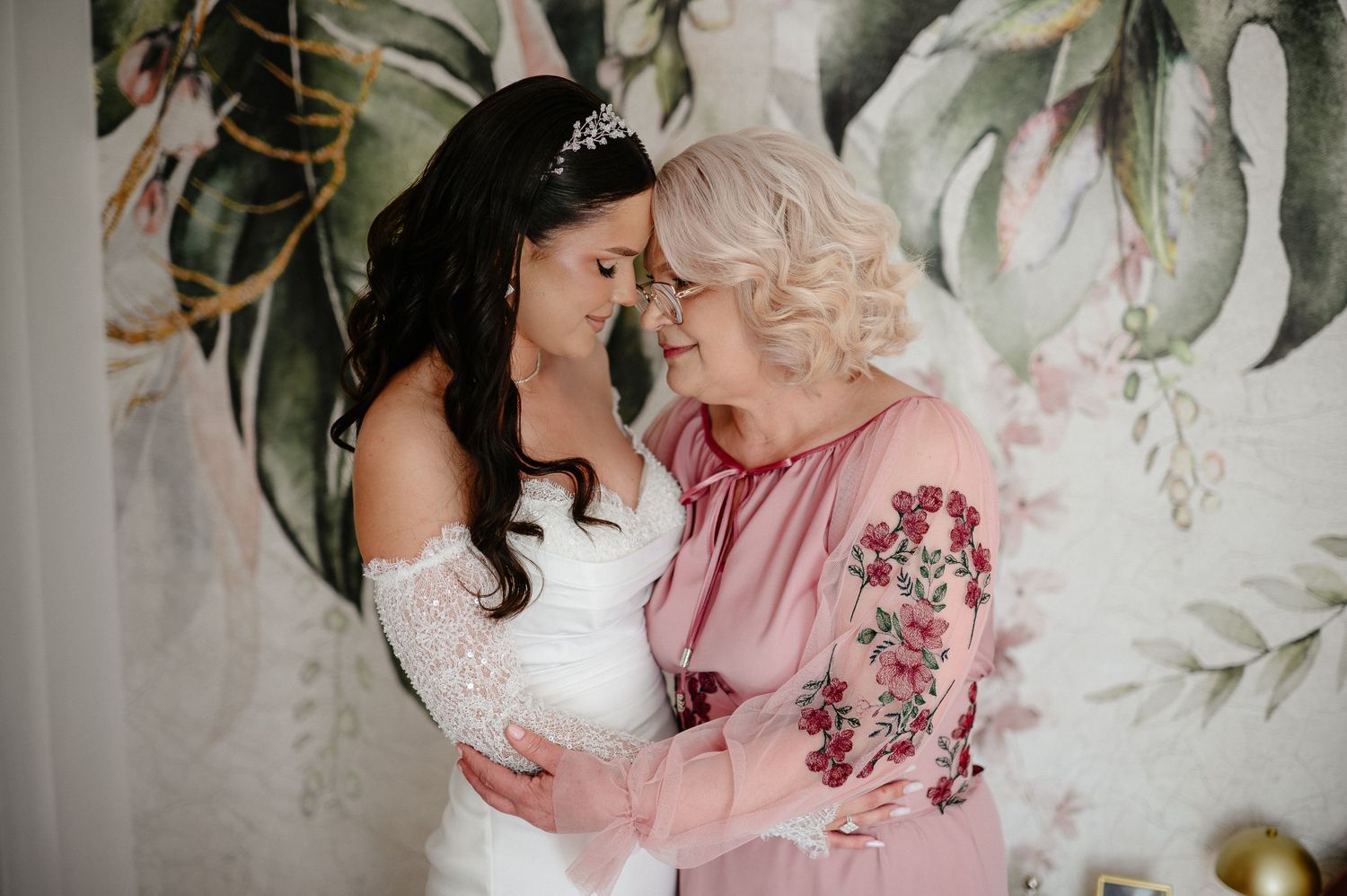 Tender moment between bride in white lace gown and mother in pink floral dress touching foreheads.