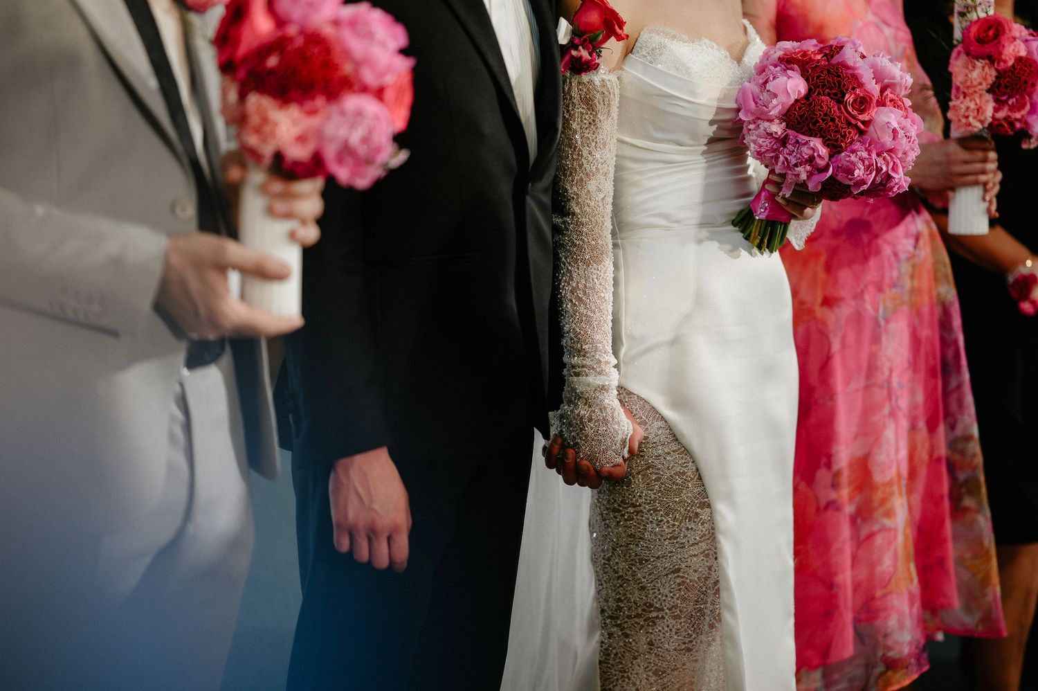 Wedding party standing in a row holding pink peony bouquets, bride in white gown with lace gloves.