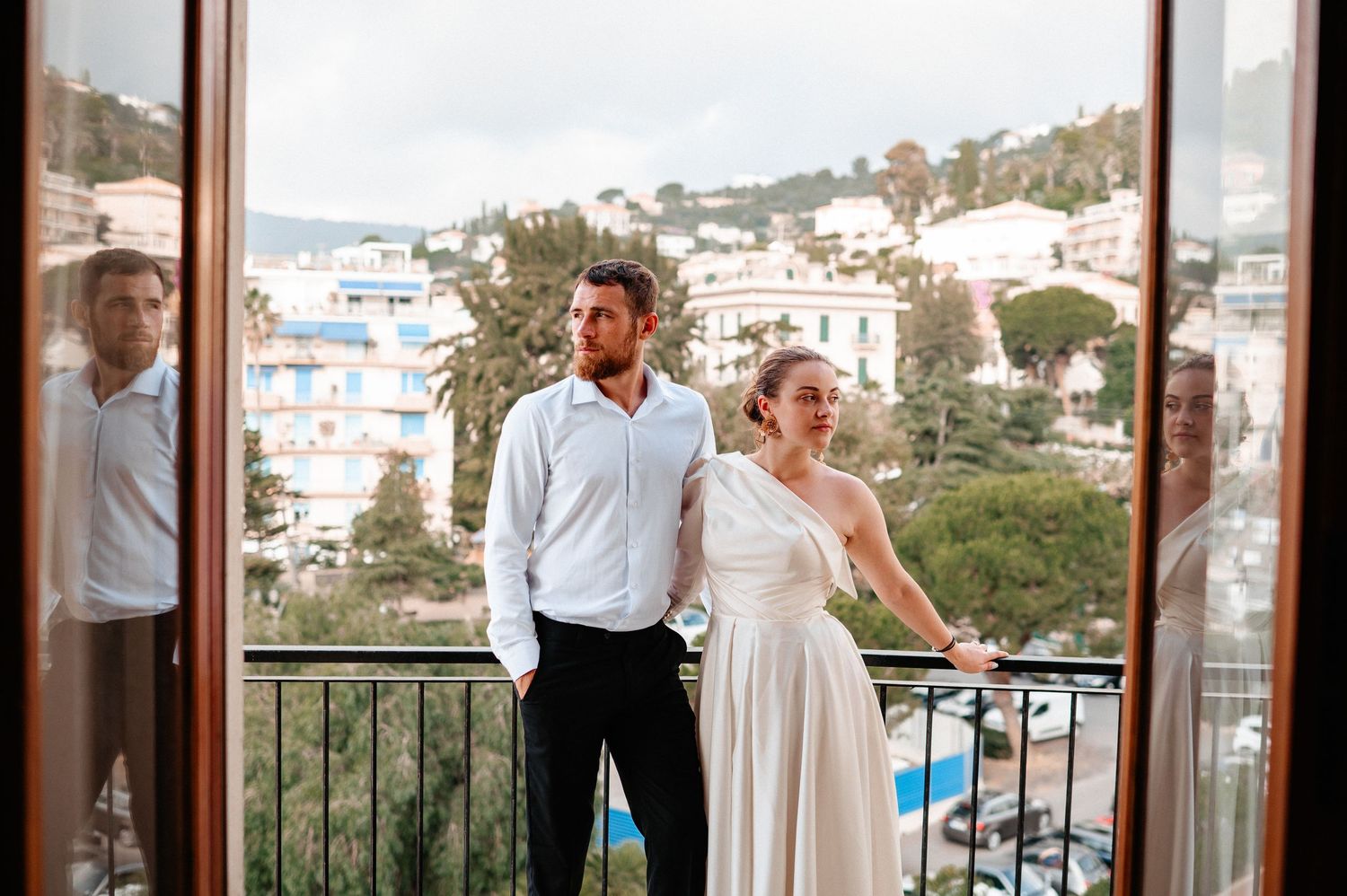 Couple in wedding attire standing on a balcony overlooking a scenic Mediterranean coastal town.