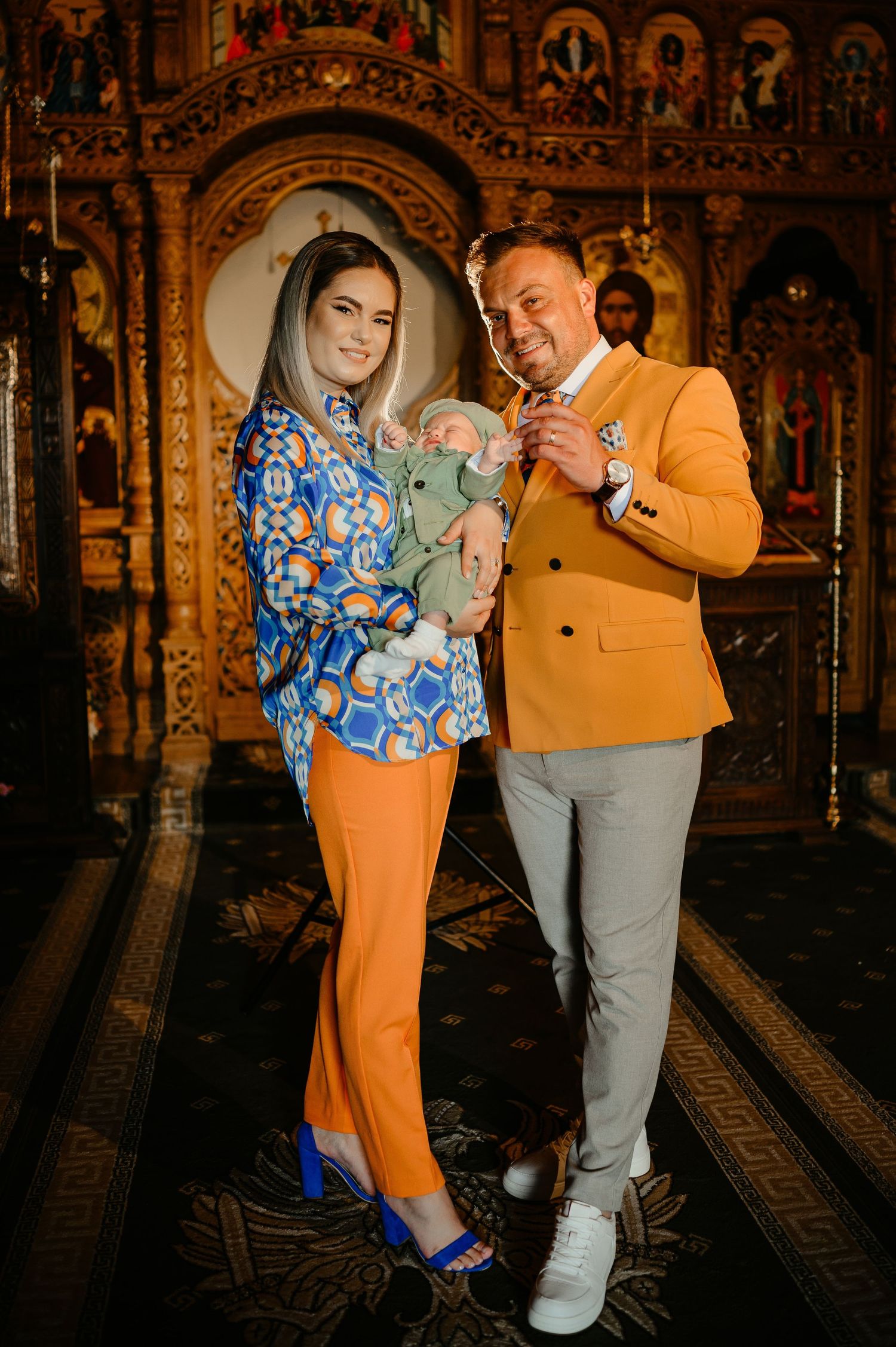 Stylish parents in orange outfits proudly holding newborn baby inside ornate Orthodox church.