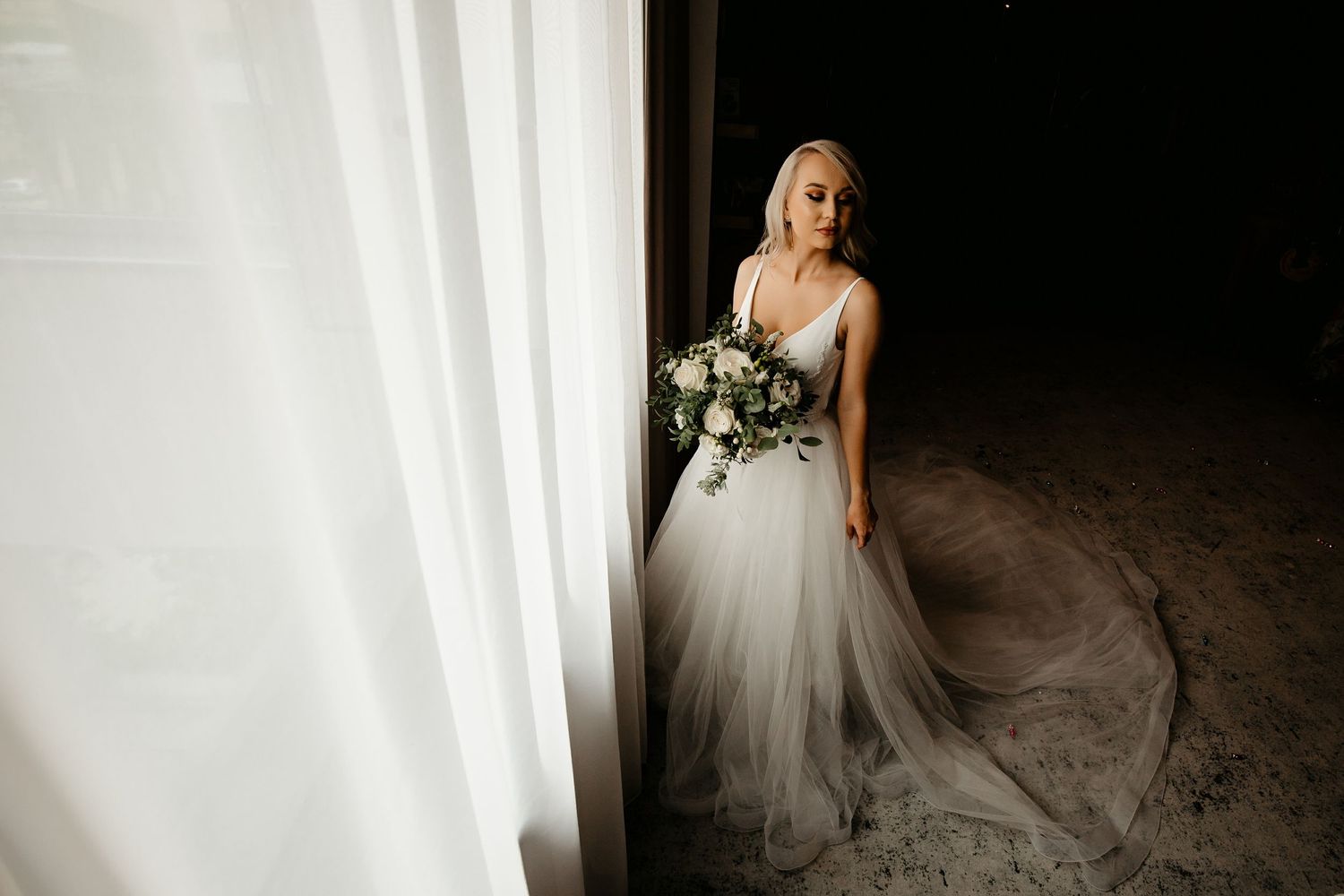 Elegant bride in flowing tulle wedding gown holding white bouquet standing in doorway with sheer curtains.