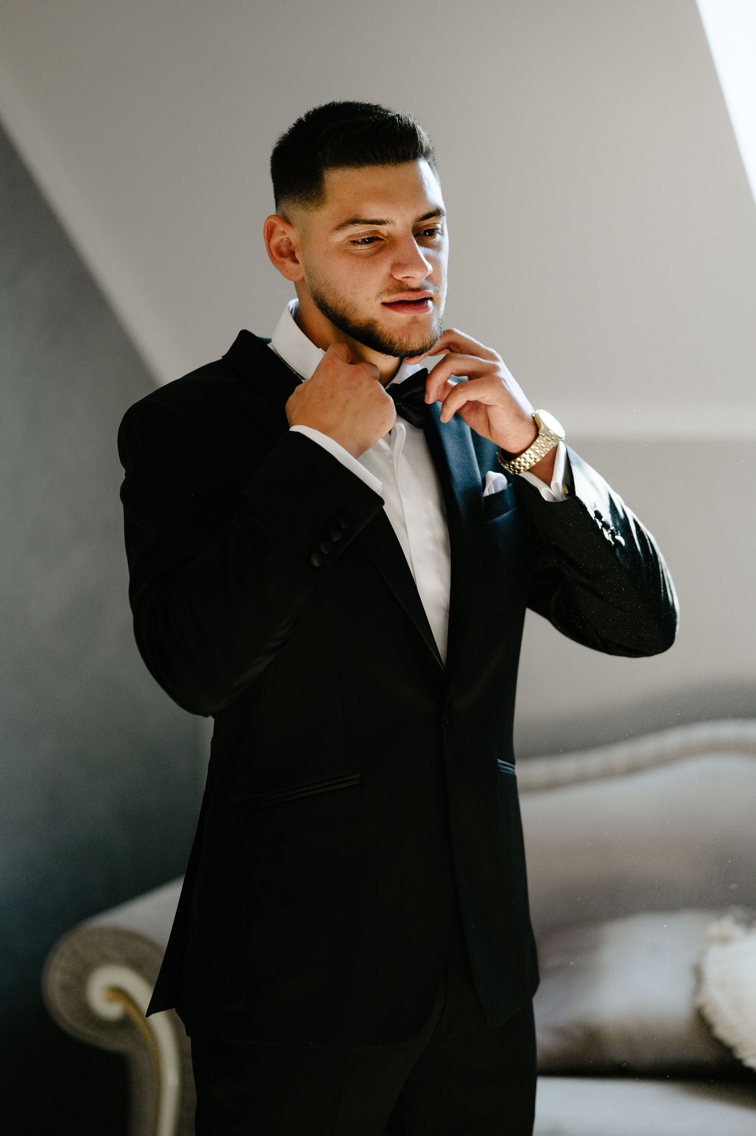 Groom in black tuxedo adjusting bow tie with gold watch in elegant room.